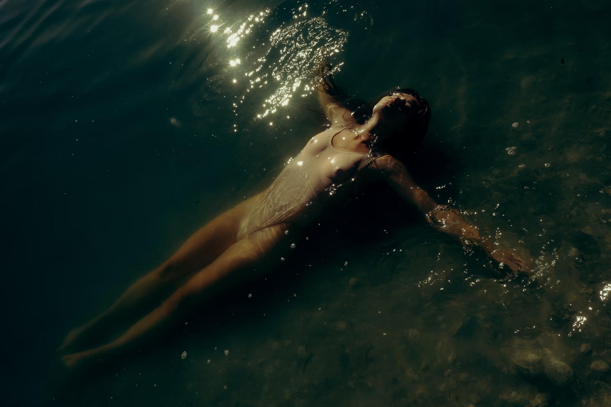 Underwater full body shot of woman swimming horizontally in dark teal lake water, graceful movement, cinematic fine art editorial