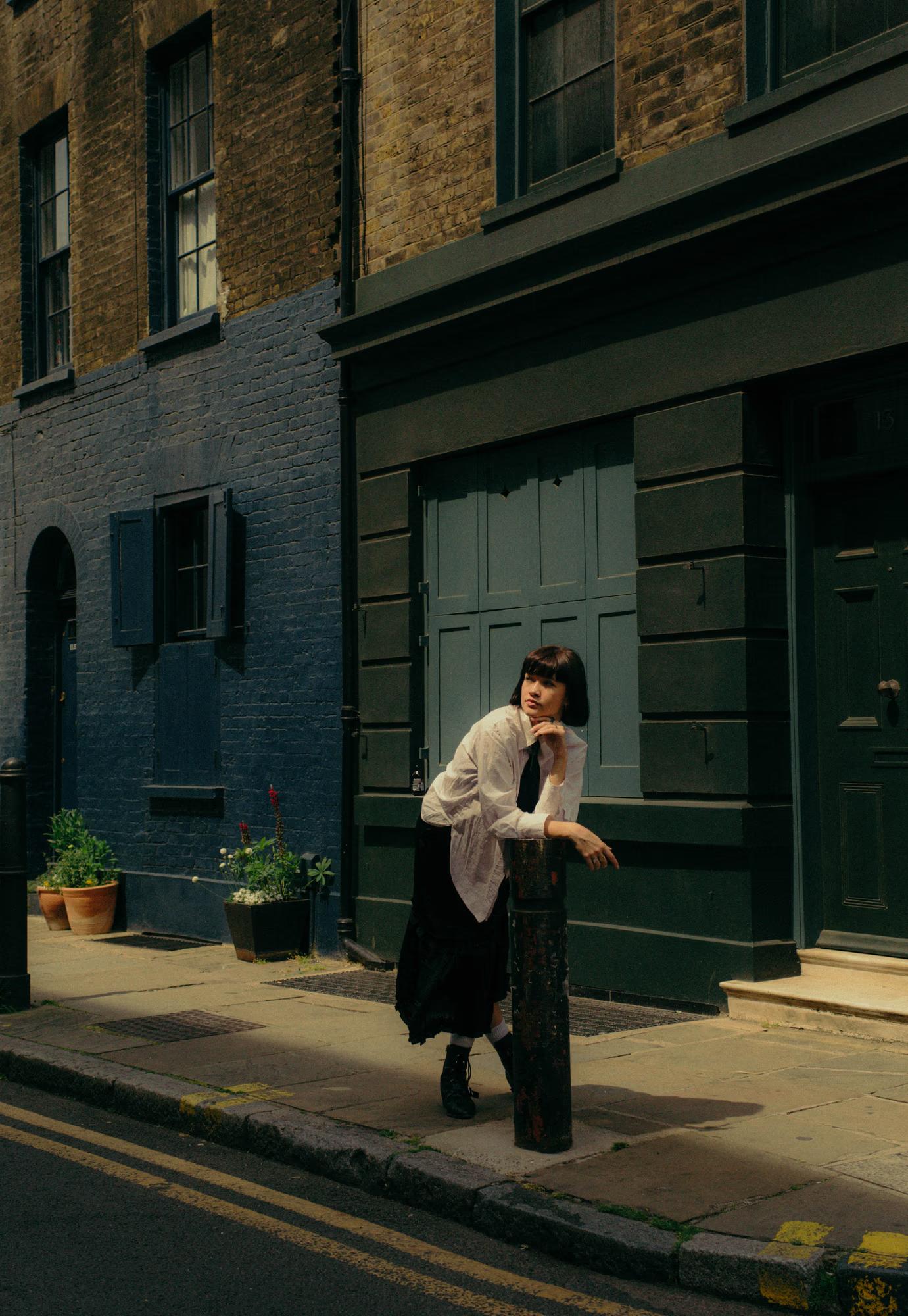 Full body fashion shot of woman in tailored white and dark outfit walking on sunlit Shoreditch London street, colorful building facades, urban street style editorial