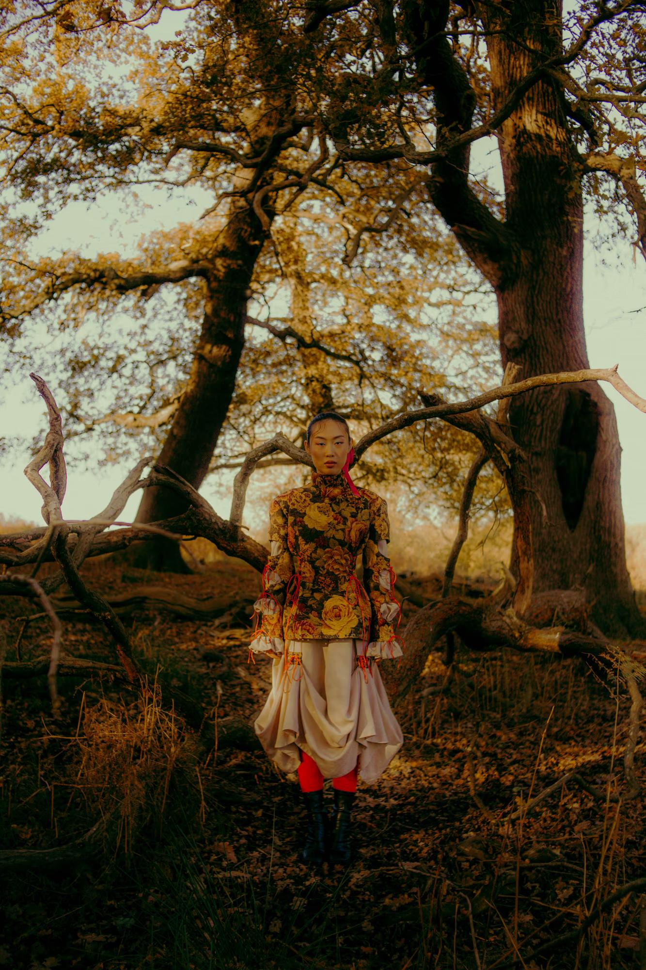 Full body fashion campaign portrait of woman in designer outfit standing beneath large oak tree, warm sunset backlight, Richmond Park London editorial
