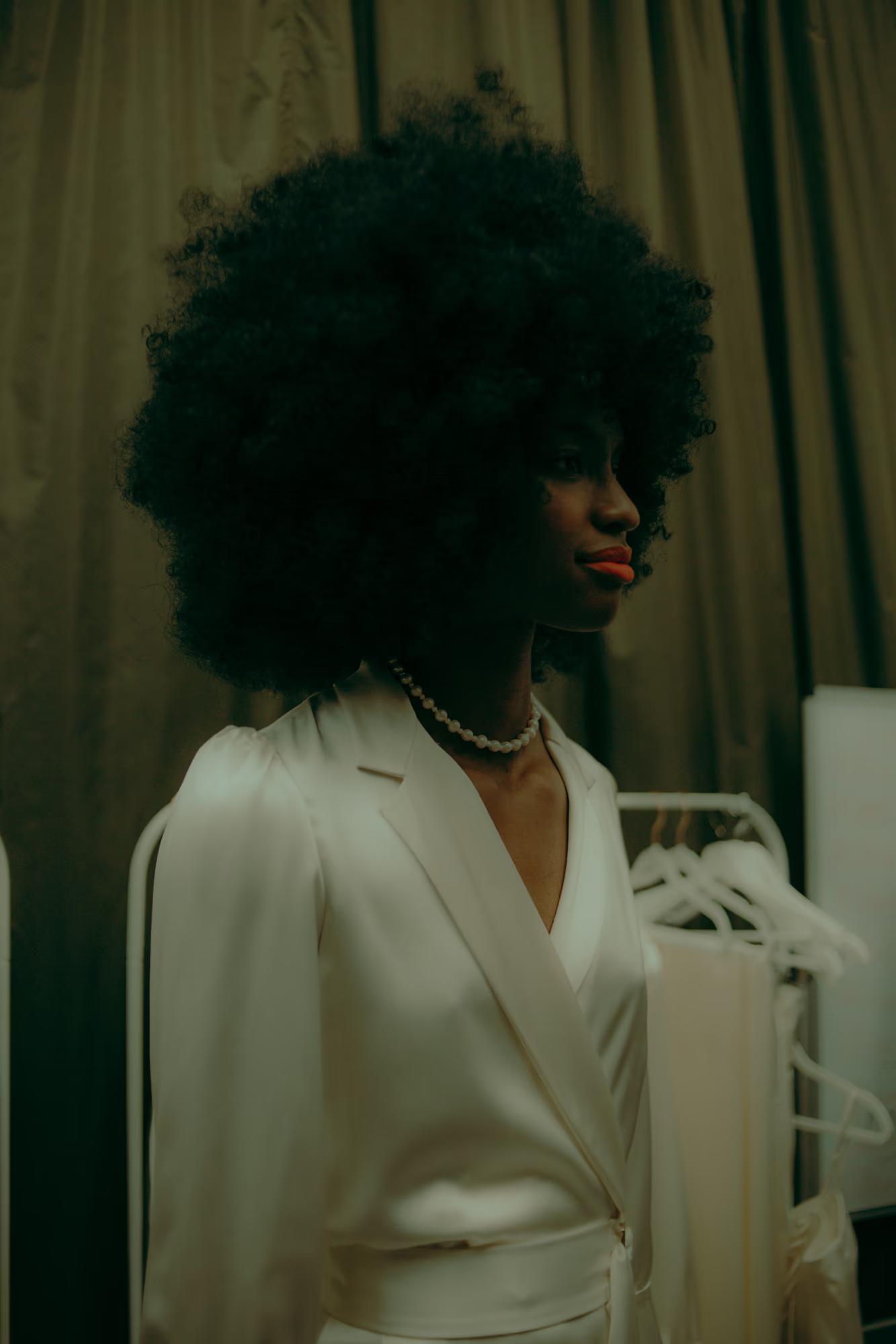 Side profile of a model with voluminous hair in a white silk robe, preparing for a London Fashion Week presentation.