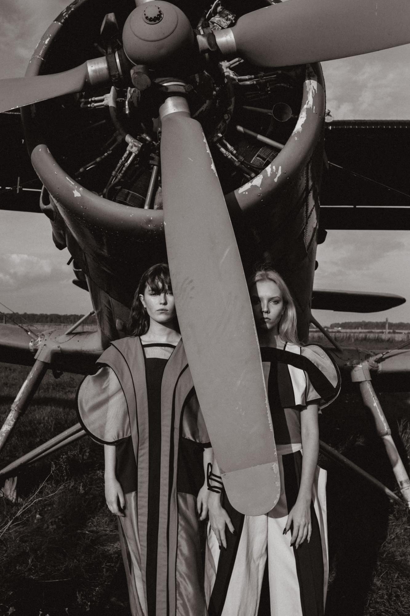  Black-and-white fashion photograph. Two models stand beneath the large radial engine and propeller of a vintage aircraft. Both wear graphic striped garments — one in a long column dress with vertical stripes, the other in a wide-shouldered black-and-white top and wide-leg trousers. Their expressions are intense and direct.