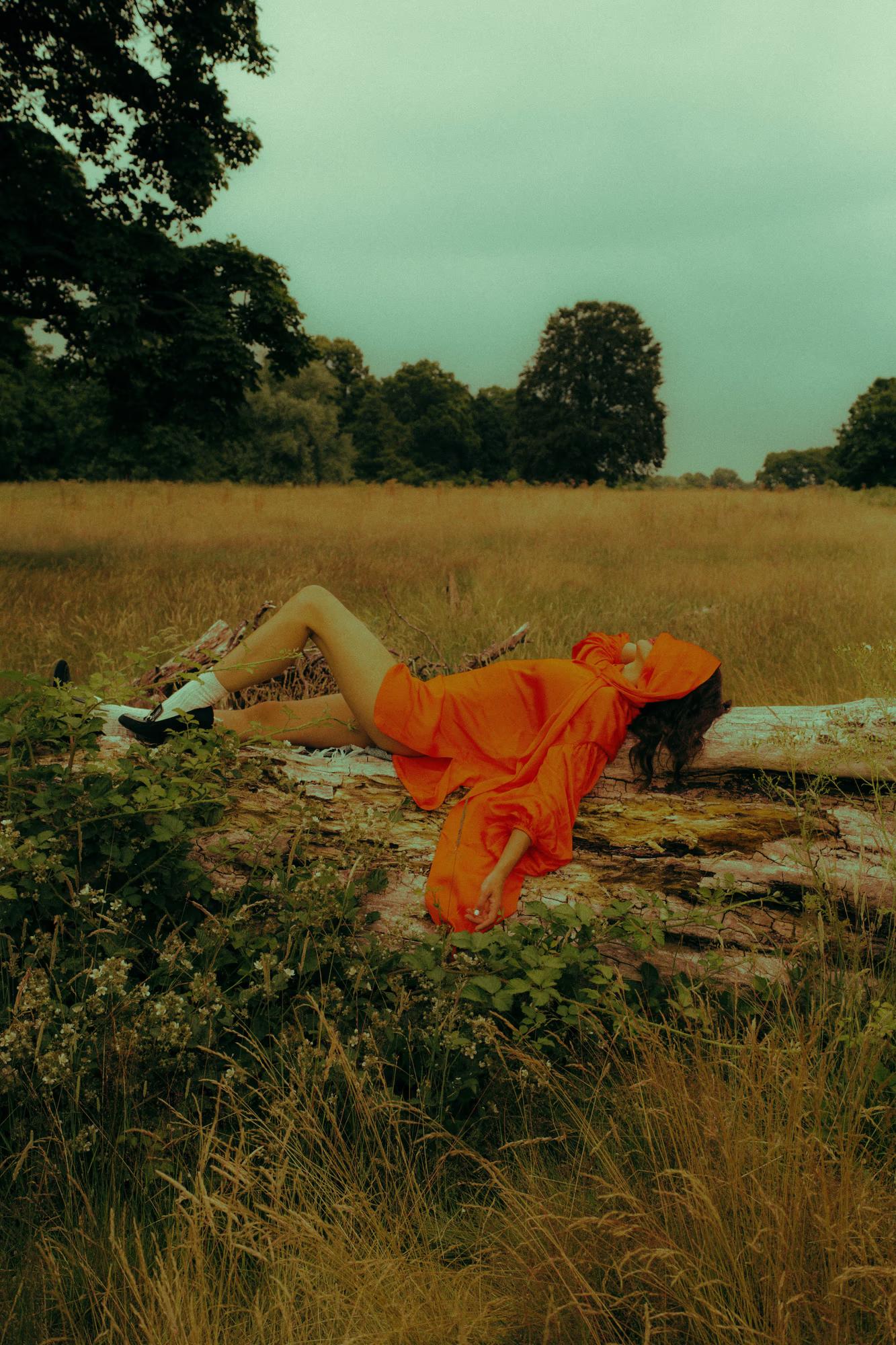  A model lies across a mossy fallen log in an open field, wearing a bright orange voluminous dress with wide sleeves. The fabric drapes over the log and spreads onto the surrounding vegetation. White socks and black shoes are visible at one end; her head is covered by the gathered fabric at the other. A stormy teal sky and distant treeline frame the scene.