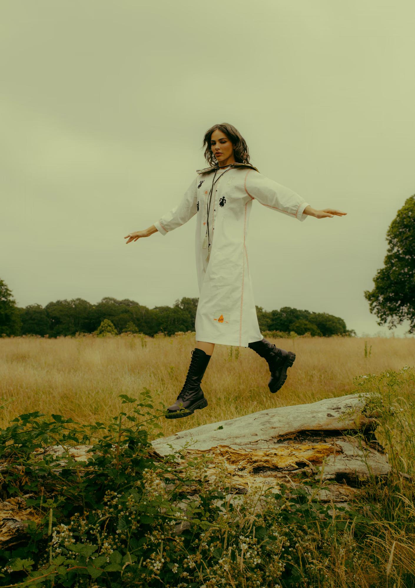 A model balances on one foot atop a fallen log in an open field, arms stretched wide to the sides. She wears a long white dress with colourful folk-inspired embroidery — small figurines and running stitch details — and tall black lace-up combat boots. The overcast sky and dry golden grass form the backdrop.
