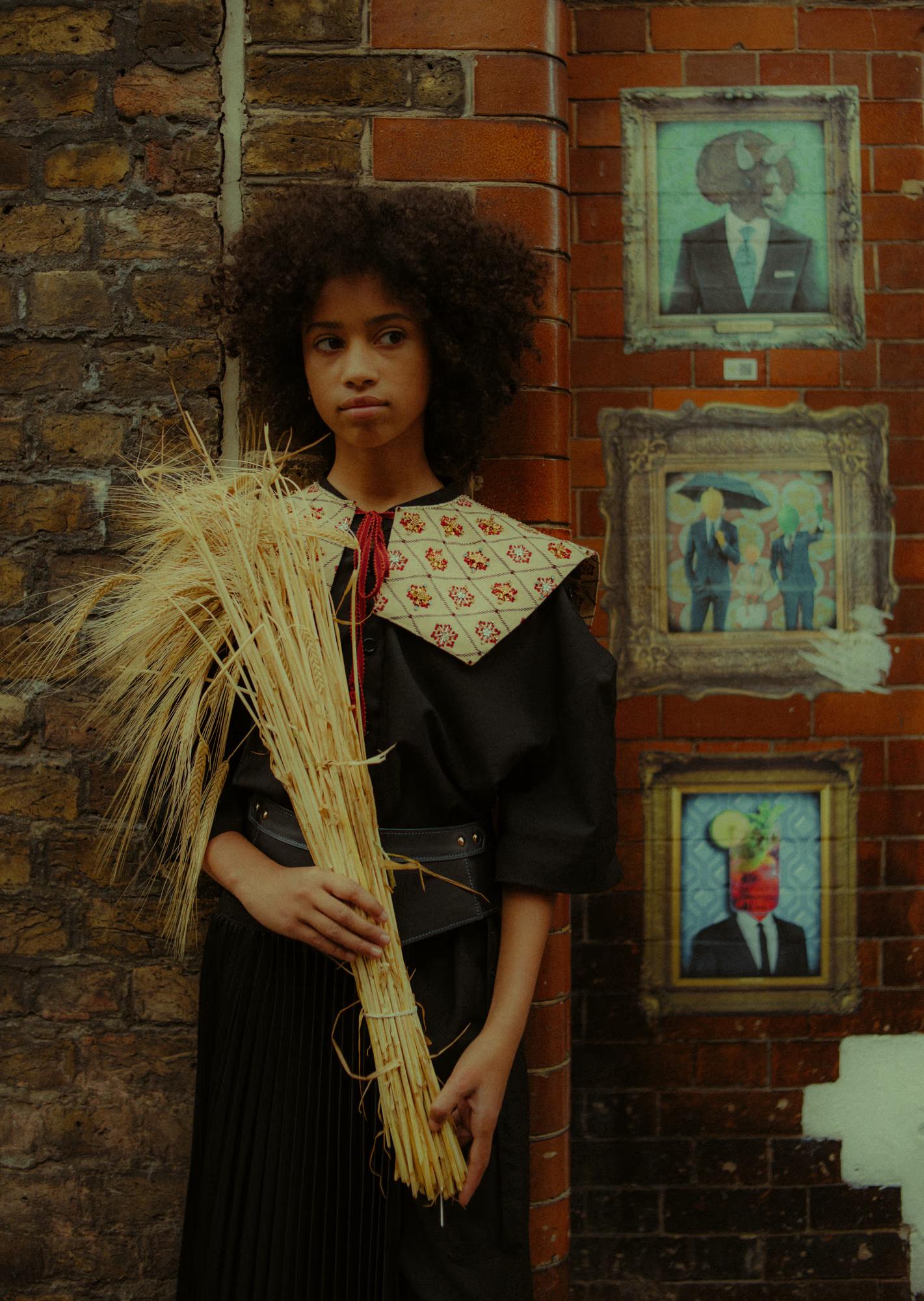 A model stands against a London brick wall holding a large bundle of dried wheat and straw, wearing a black dress with a wide embroidered diamond-grid collar with red floral motifs, a leather belt, and a black pleated skirt. Three framed surrealist oil paintings — depicting figures with animal or object heads in formal attire — hang on the brick wall behind her.