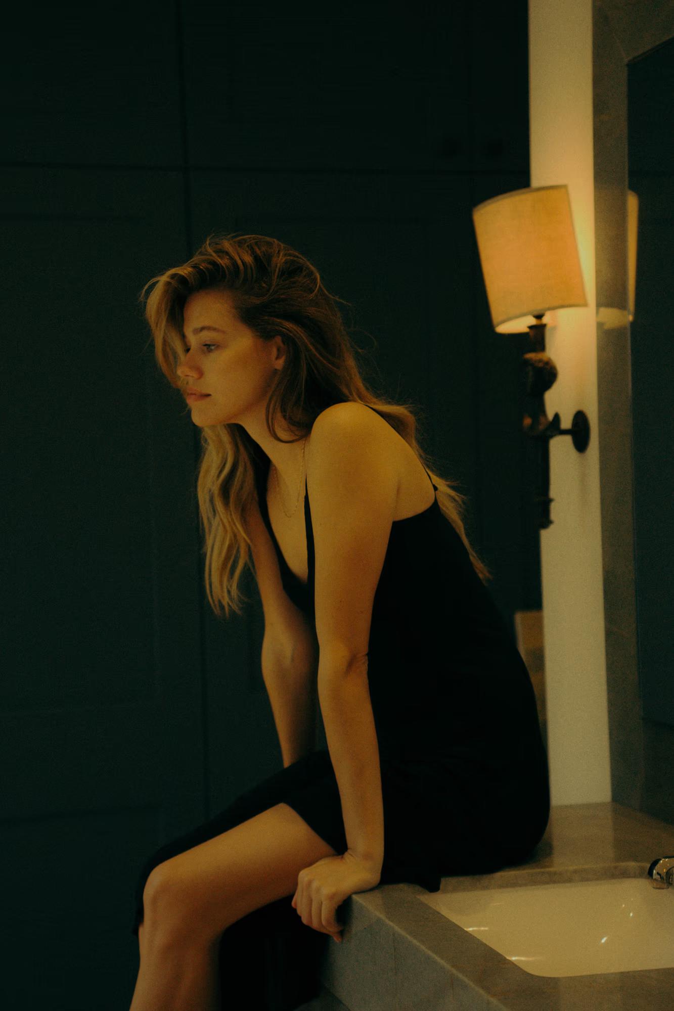 A model sits hunched on the edge of a bathroom surface in a dark room, wearing a black spaghetti-strap slip dress. She looks downward, hair falling forward, lit only by the warm glow of a wall sconce lamp to her right. The mood is quiet and introspective.