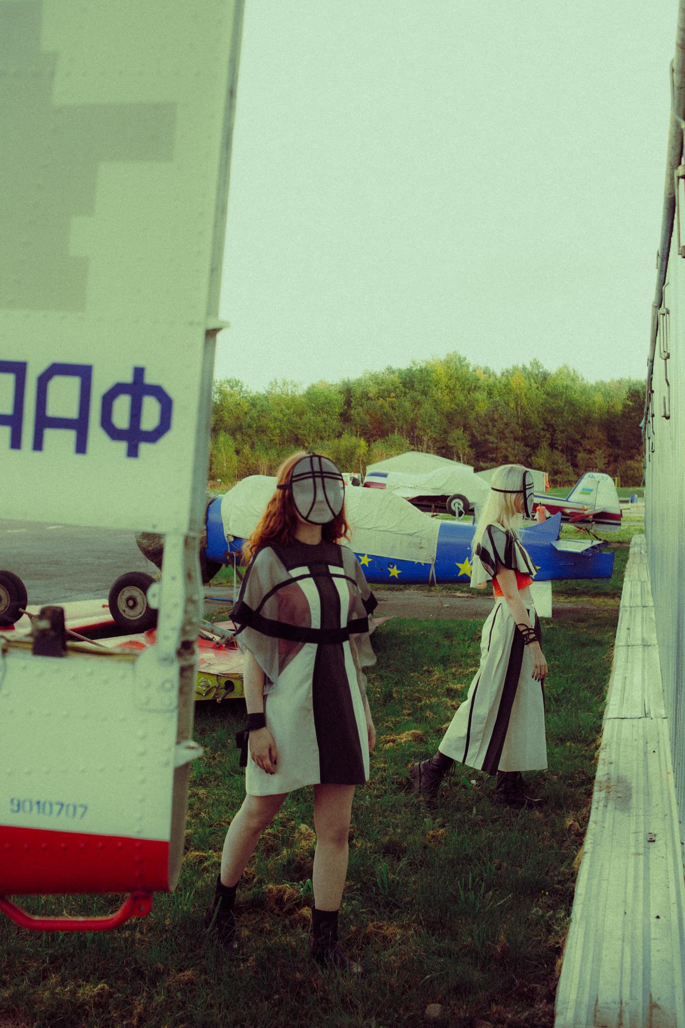 Two models stand between the tail sections of two parked aircraft on a grass airfield. The foreground model wears a sheer grid mask and a graphic black-and-white mini dress; the second model, facing away, wears a long black-and-white striped coat. Cyrillic lettering "ААФ" is visible on the plane to the left.