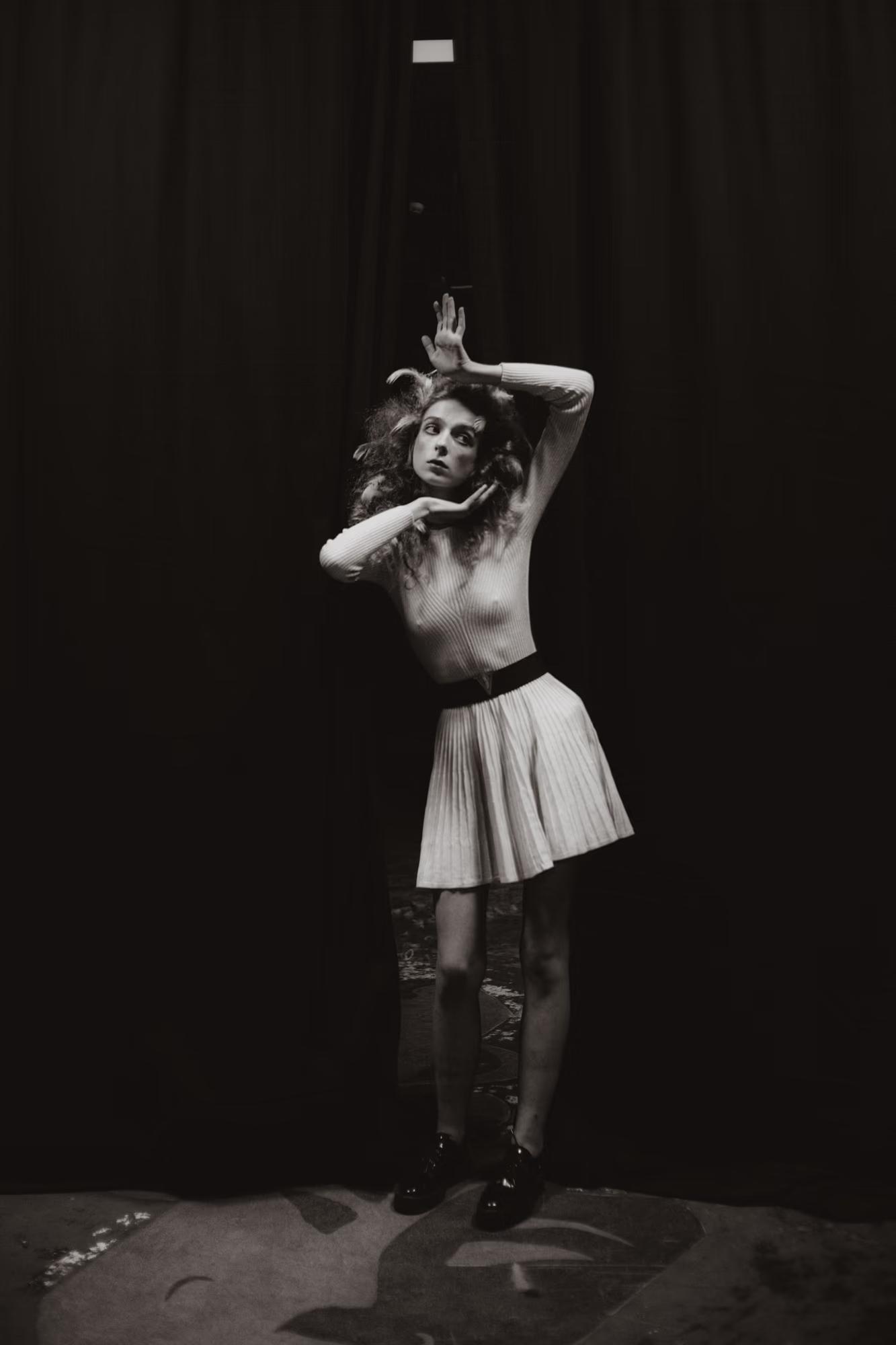 Artistic full-body pose of a model in a white mini dress on a theater stage.