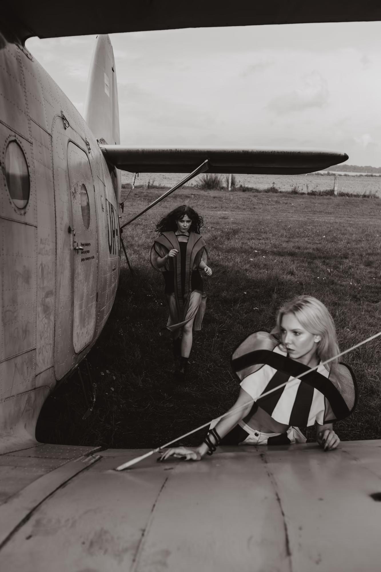  Black-and-white photograph. A blonde model lies draped across the top surface of an aircraft wing in a bold geometric striped top, one hand on the wing surface. In the background, a dark-haired model in a structured vest sprints across the grass behind the plane's fuselage.