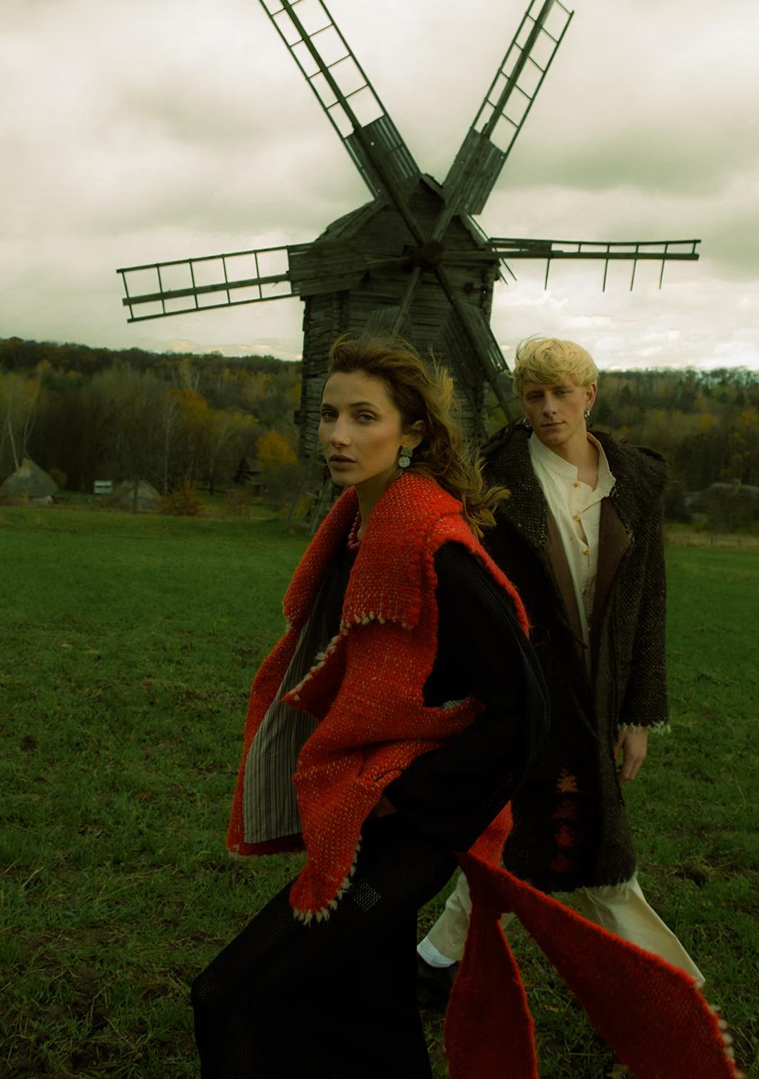 A model in a dramatic red cloak walking past a historic wooden windmill at the Pirogovo ethno-park, Kyiv.