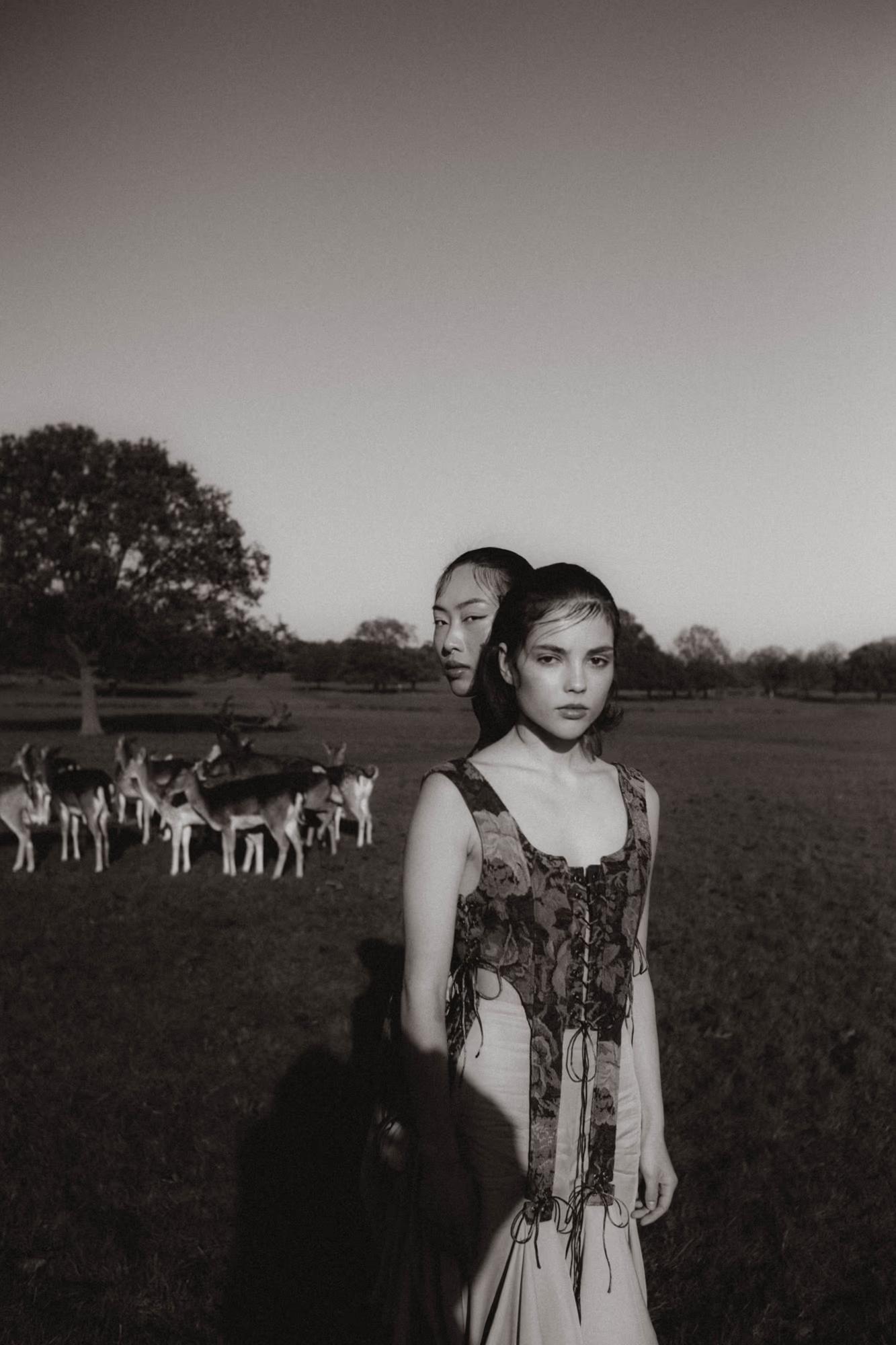 Black and white full body fashion shot of woman in flowing designer dress standing in Richmond Park London field with deer grazing in background