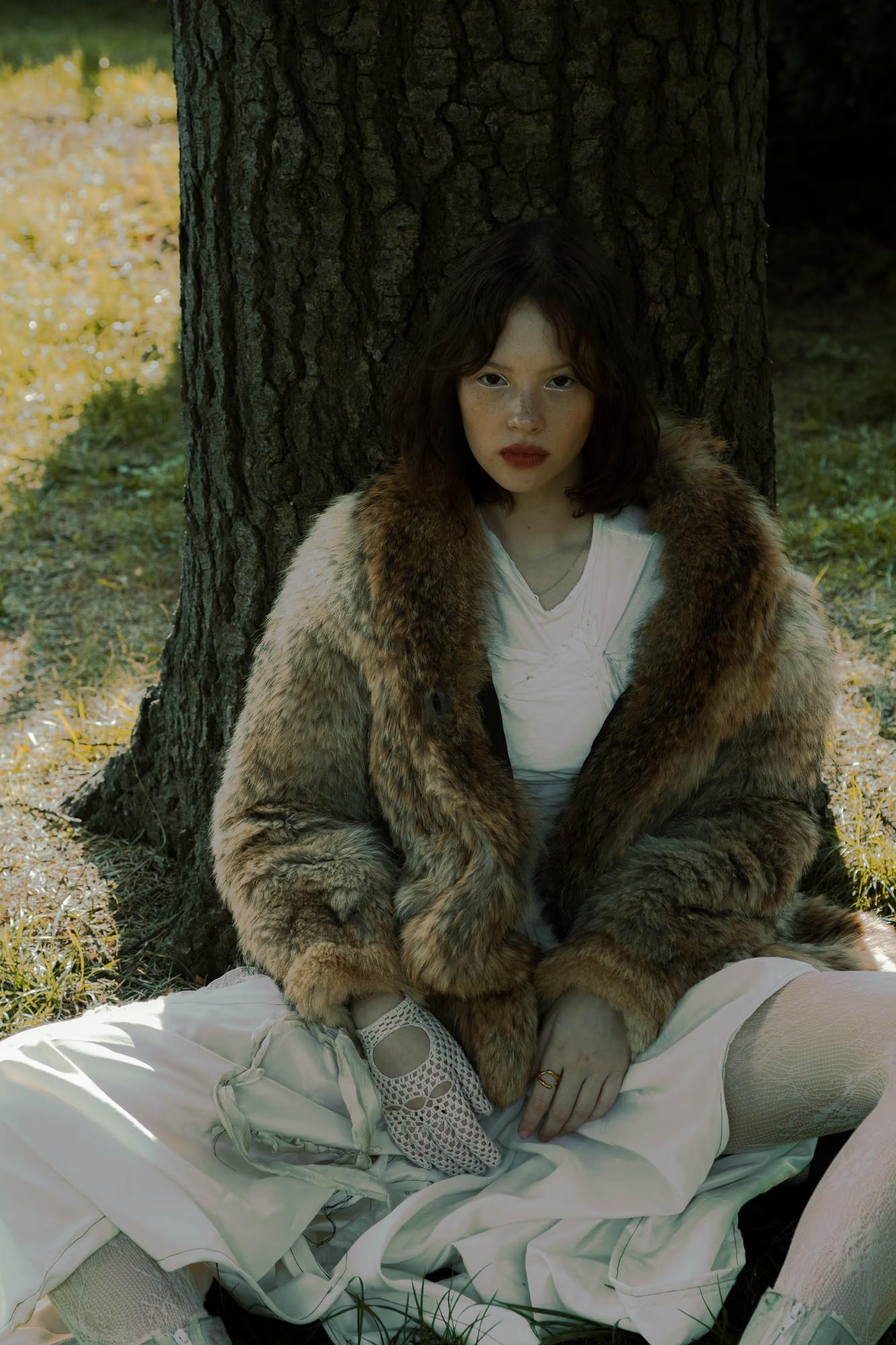 Full body fashion art shot of woman in fur and light outfit seated in dark forest, mysterious atmospheric editorial