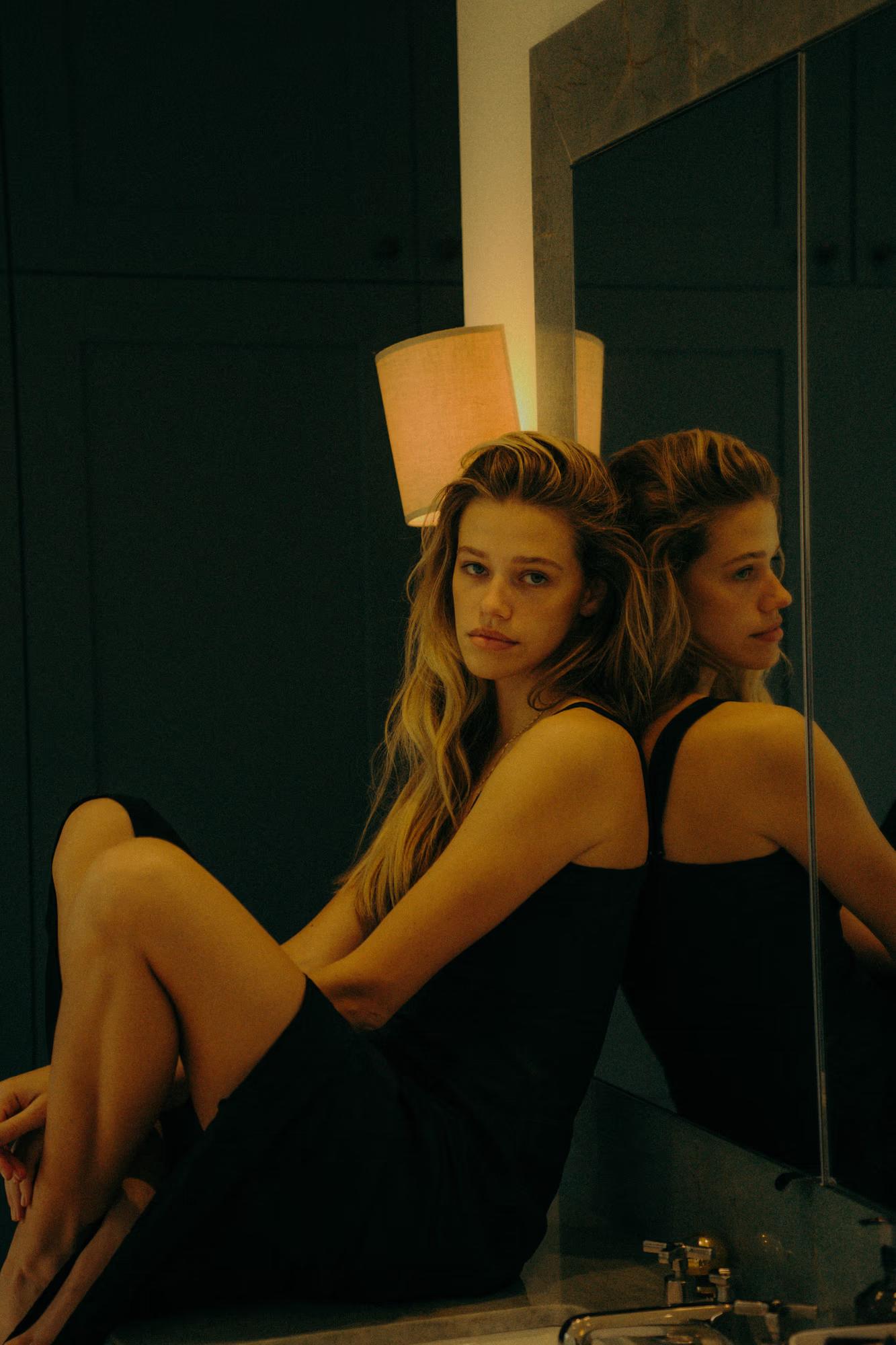 A model sits on a bathroom counter with her back against a large mirror, legs folded, wearing a black sleeveless slip dress. Her reflection is visible in the mirror behind her, and a glowing cylindrical wall lamp is centred above. The dark teal panelled wall and a small basin tap are visible in the foreground. She looks at the camera with a calm, measured expression.