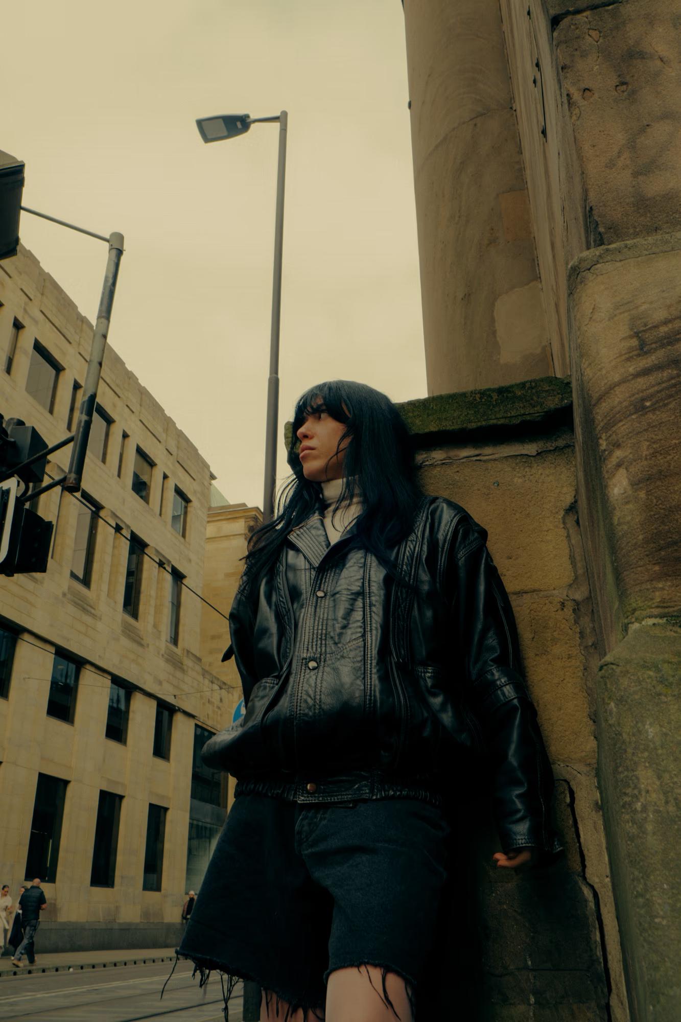 Full body fashion shot of person in dark oversized jacket crouching on Manchester pavement with urban architecture backdrop, gritty street style editorial