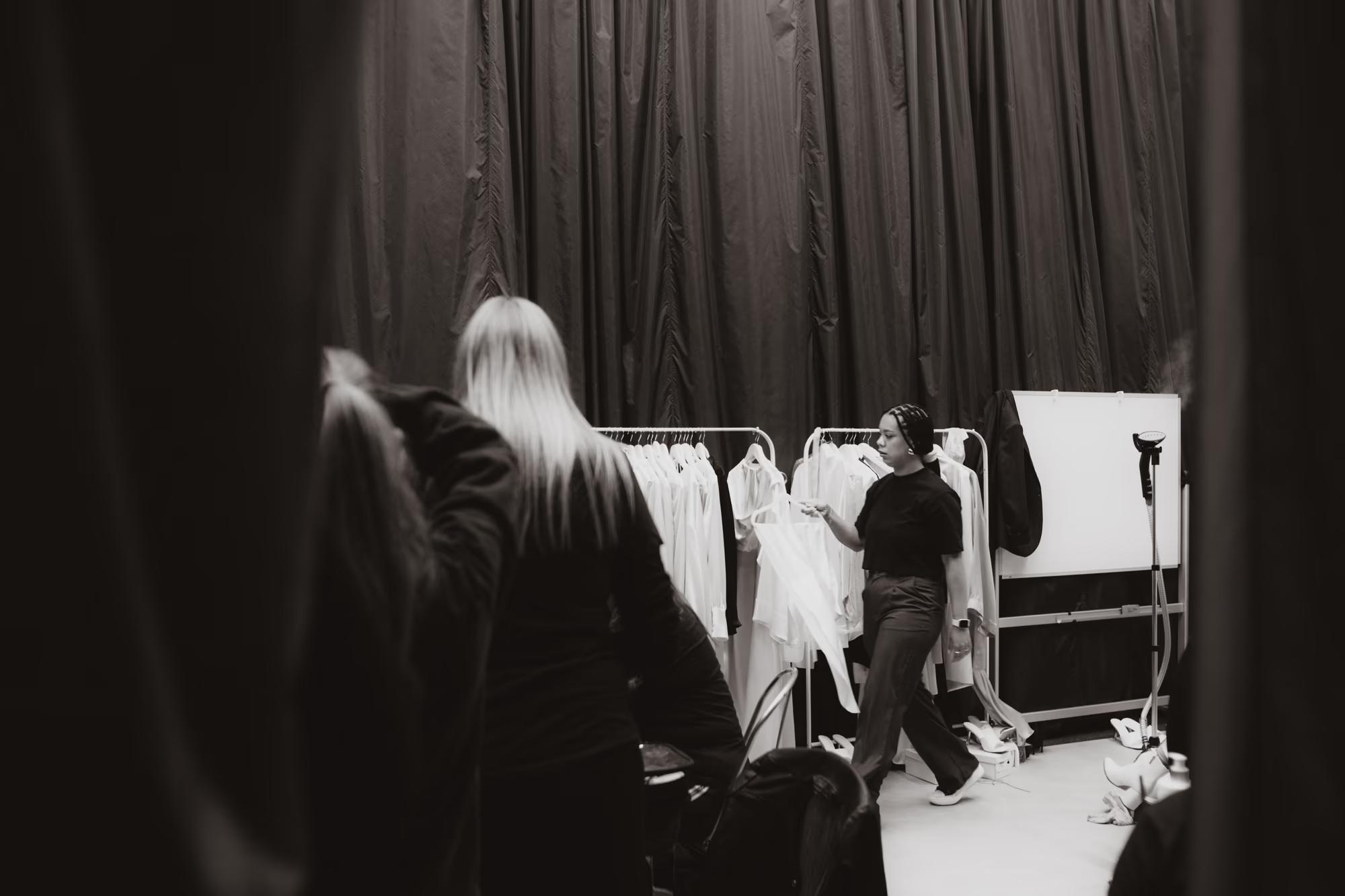 Wide cinematic shot looking toward the runway from behind the dark stage curtains, showing the hustle of production.