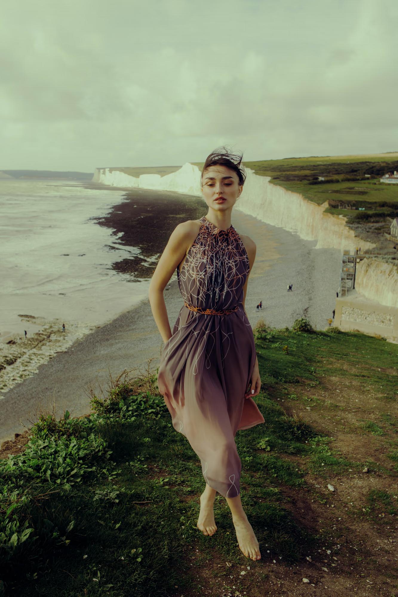Full body fashion campaign shot of woman in purple designer dress walking on Eastbourne beach shoreline, soft coastal light, luxury fashion editorial