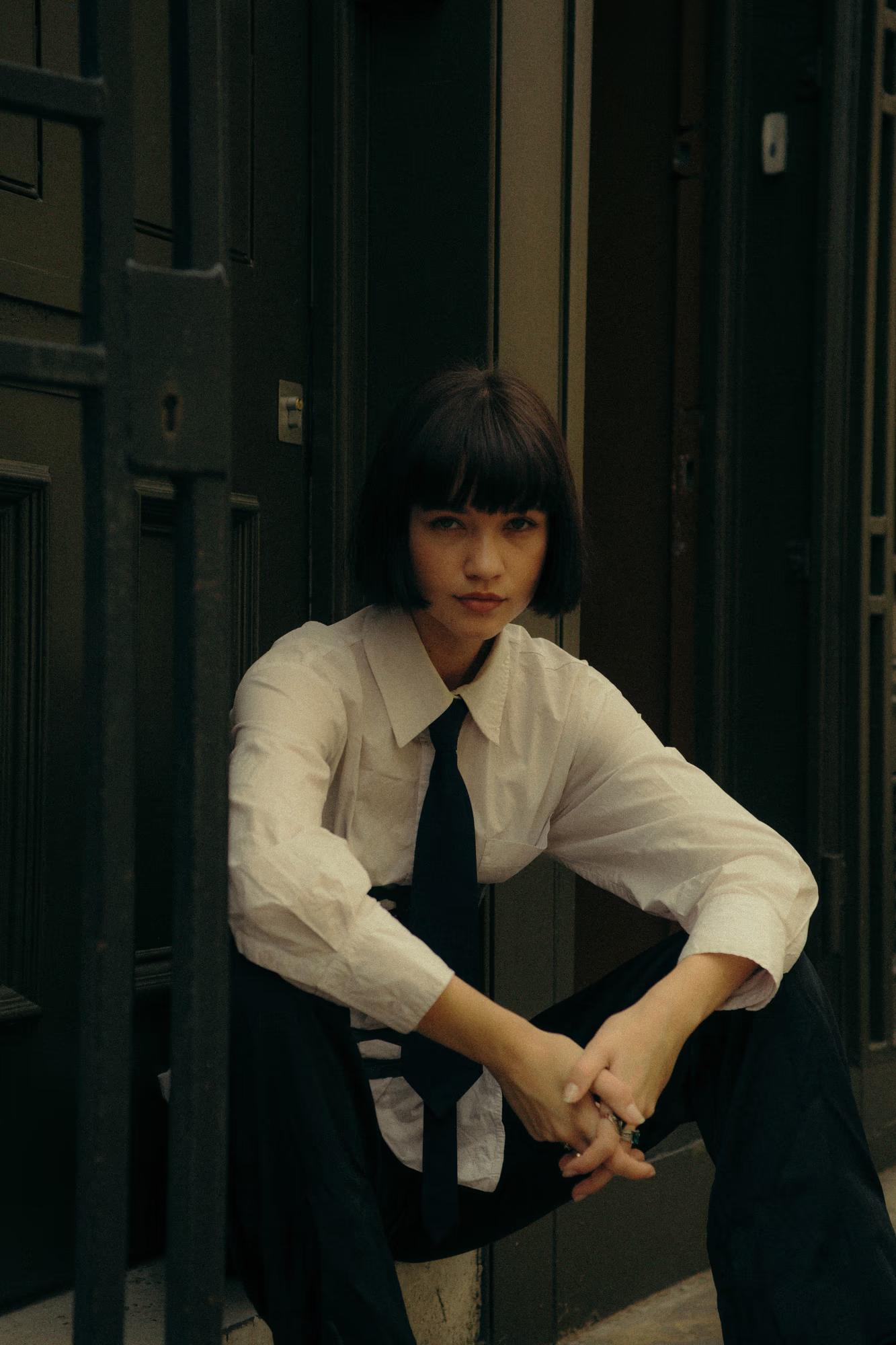 Medium fashion shot of woman in white shirt and dark tie seated on steps, androgynous tailored style, moody cinematic tones, Shoreditch London