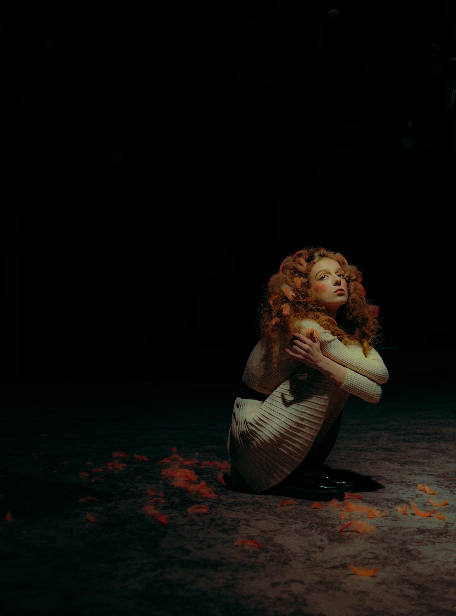 Moody shot of a woman crouching on a dark stage, high-contrast theatrical lighting.
