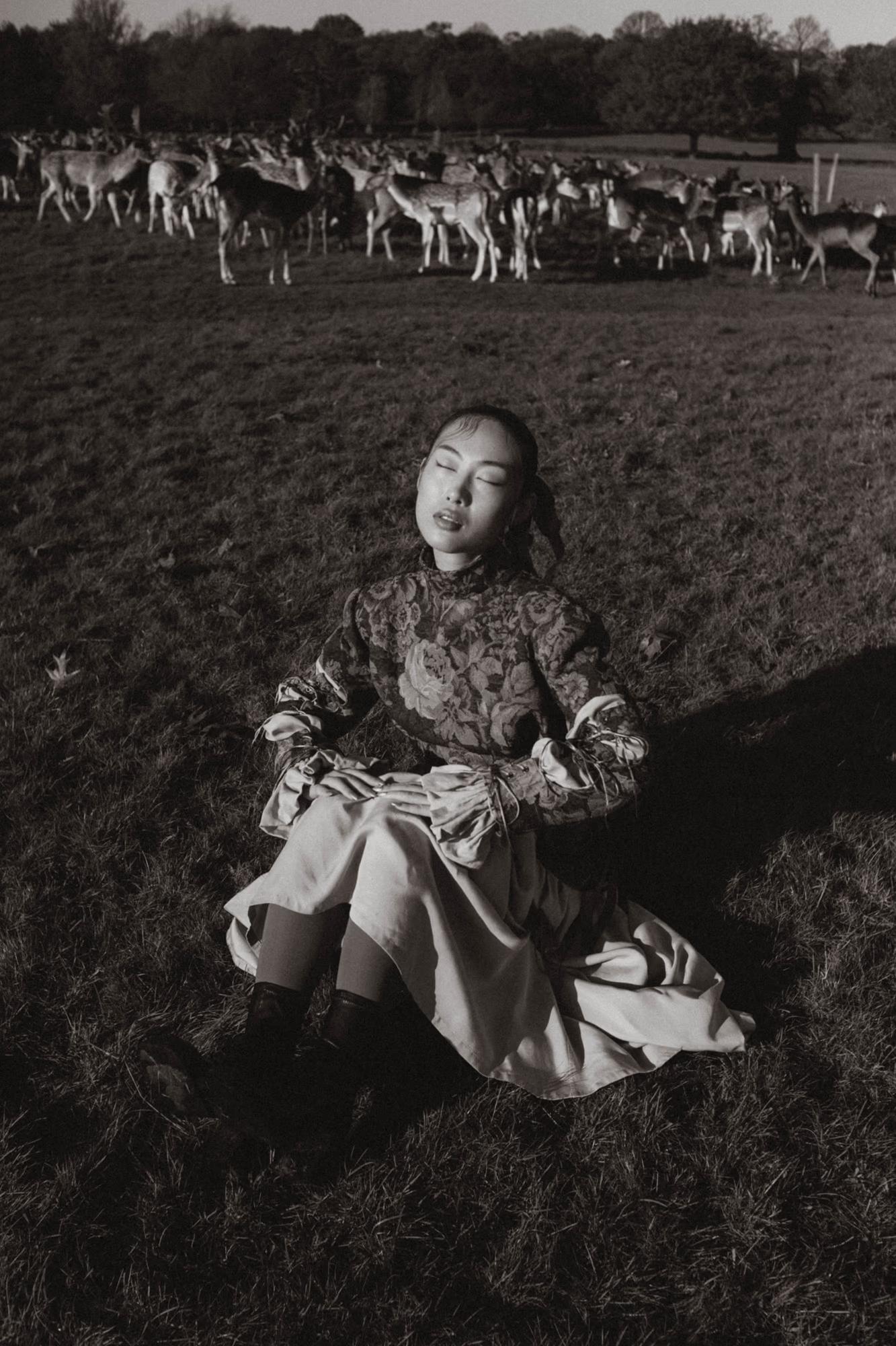 Black and white full body fashion shot of woman in designer dress seated in Richmond Park London field with deer grazing in background, fine art editorial