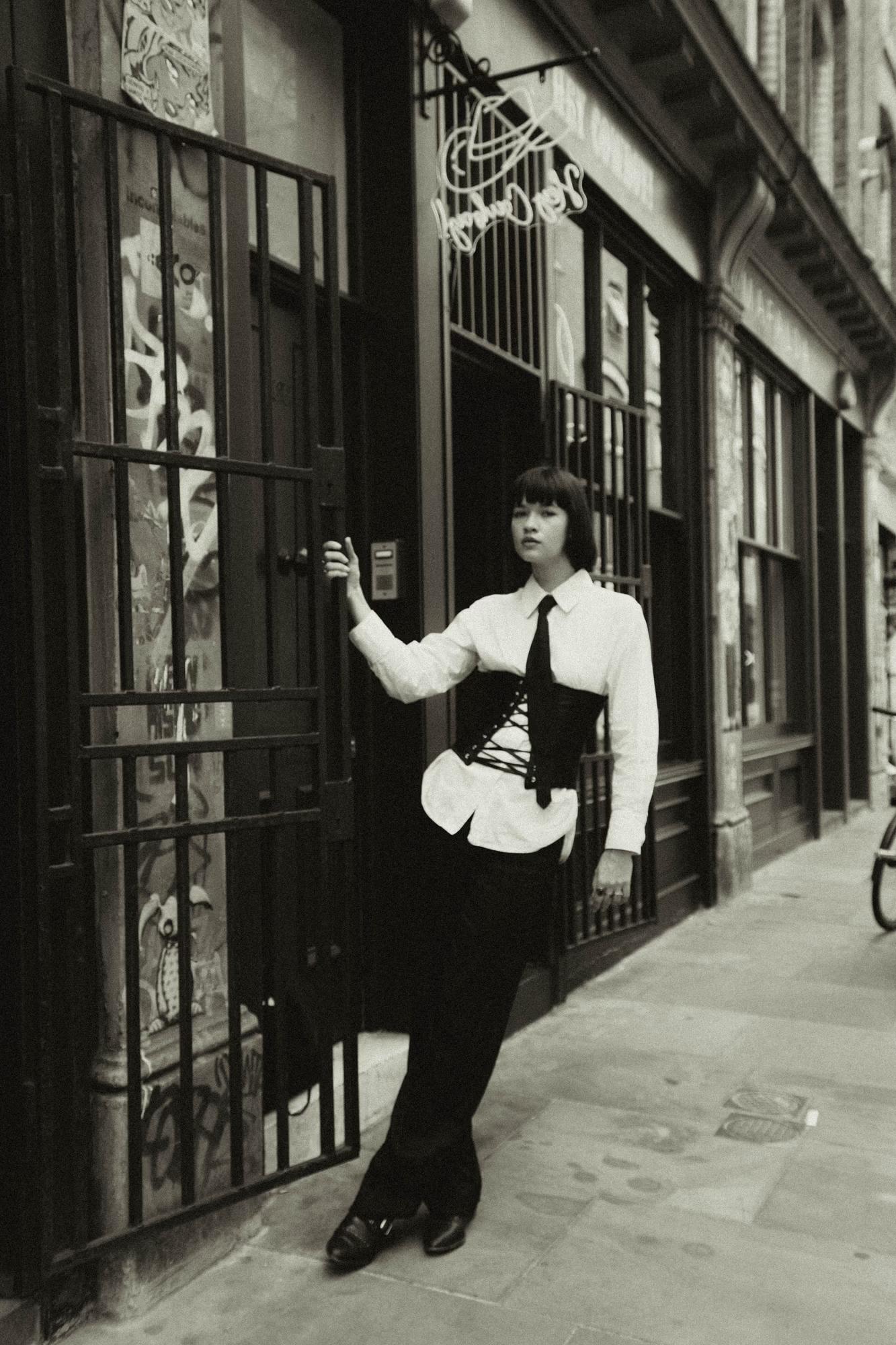 Full body fashion shot of woman in dark tailored outfit leaning against classic Shoreditch London brick building, moody urban street style editorial