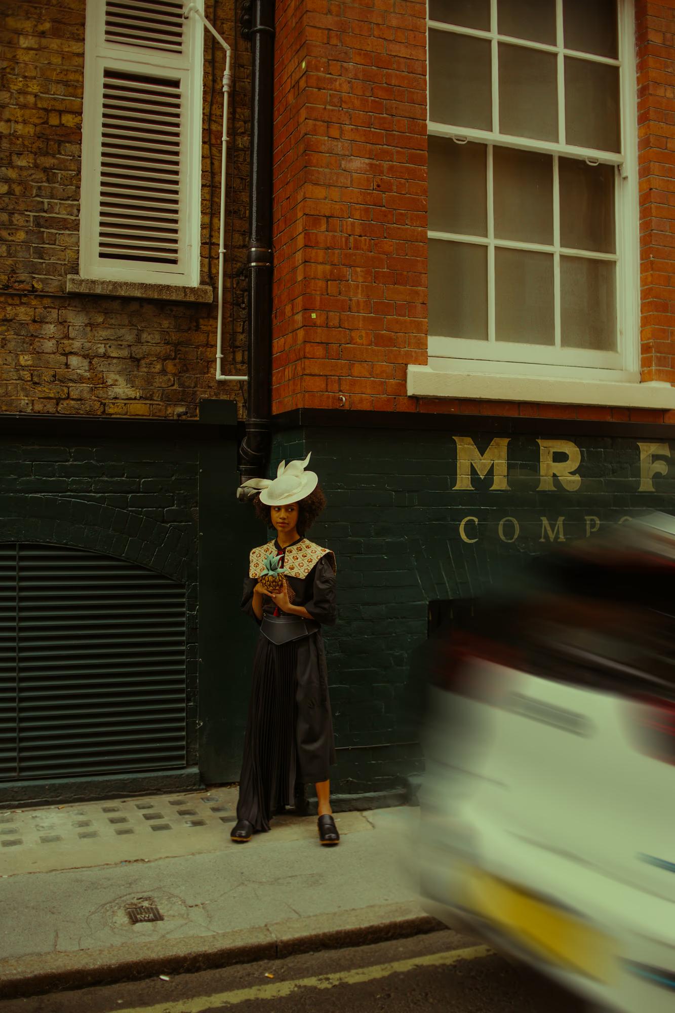 A model stands on a narrow London pavement beside a black drainpipe, wearing a black pleated midi skirt, an embroidered patterned collar top, a wide leather belt, and a sculptural white fascinator hat with a feather. A car blurs past in the foreground. A dark green shopfront with gold lettering is visible behind her.