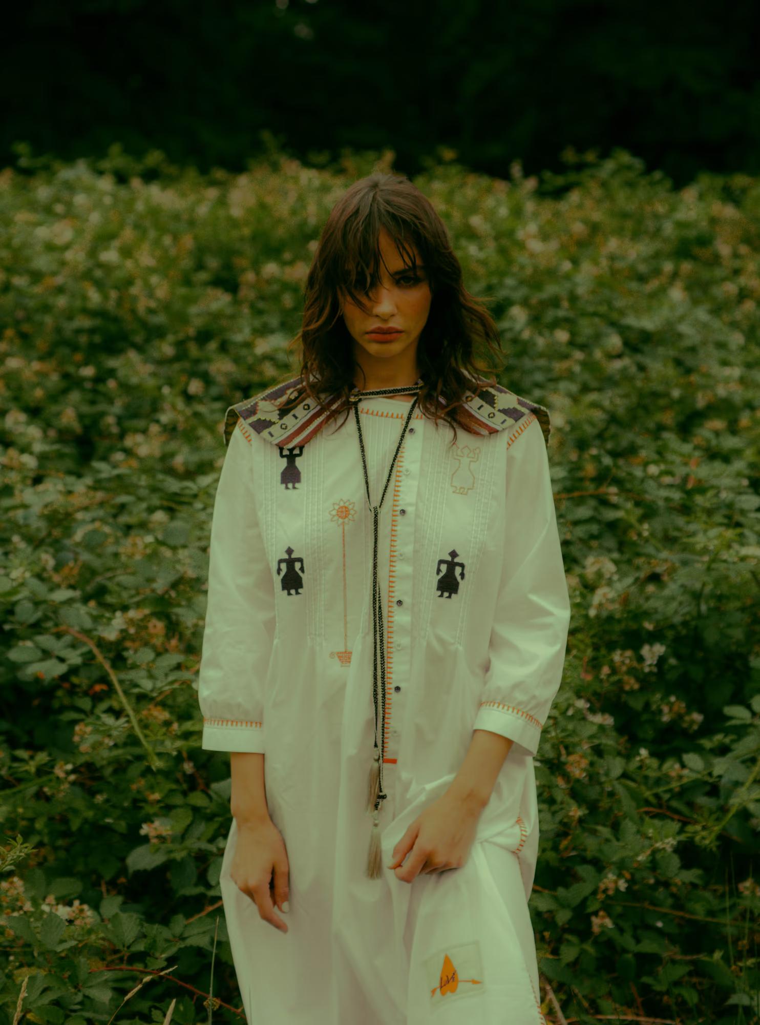  A model stands amid wild greenery and white wildflowers, wearing a white long-sleeved dress covered in folk embroidery — small human figurines, geometric border details in orange and black, and a coloured yoke. A long braided tassel necklace hangs at the centre. She faces the camera with a direct, composed expression.