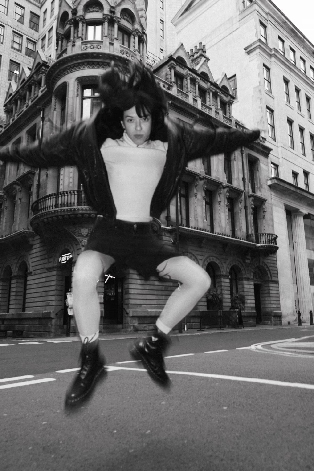 Full body black and white fashion shot of person in dark streetwear jumping or posing dynamically on Manchester street, urban editorial photography