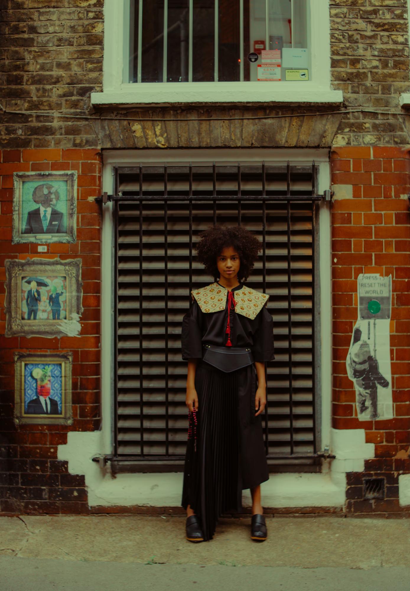 A model stands in front of a metal-shuttered doorway on a London brick building, wearing a black blouse with a wide embroidered folk-style collar, a red tassel tie, a structured leather belt, and a black pleated skirt. Ornately framed surrealist portrait paintings are affixed to the brick wall to her left. A street sticker reading "Press to Reset the World" is visible to her right.