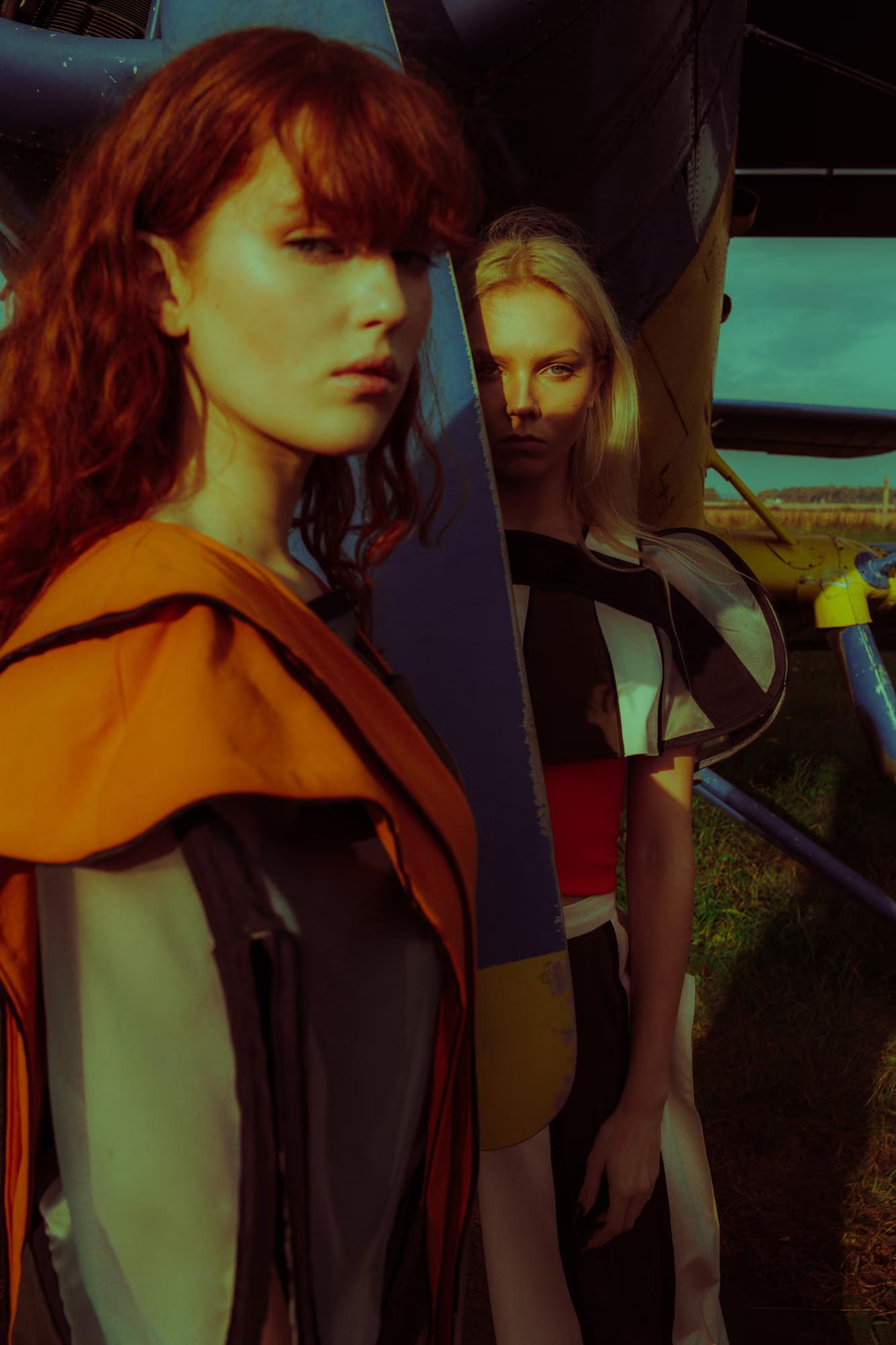 Two models posing close together against the fuselage of a vintage aircraft. The foreground model has long red hair and wears a draped amber-and-charcoal outfit; the background model is blonde and wears a geometric black, white, and red structured top. Warm, moody lighting with a teal sky.
