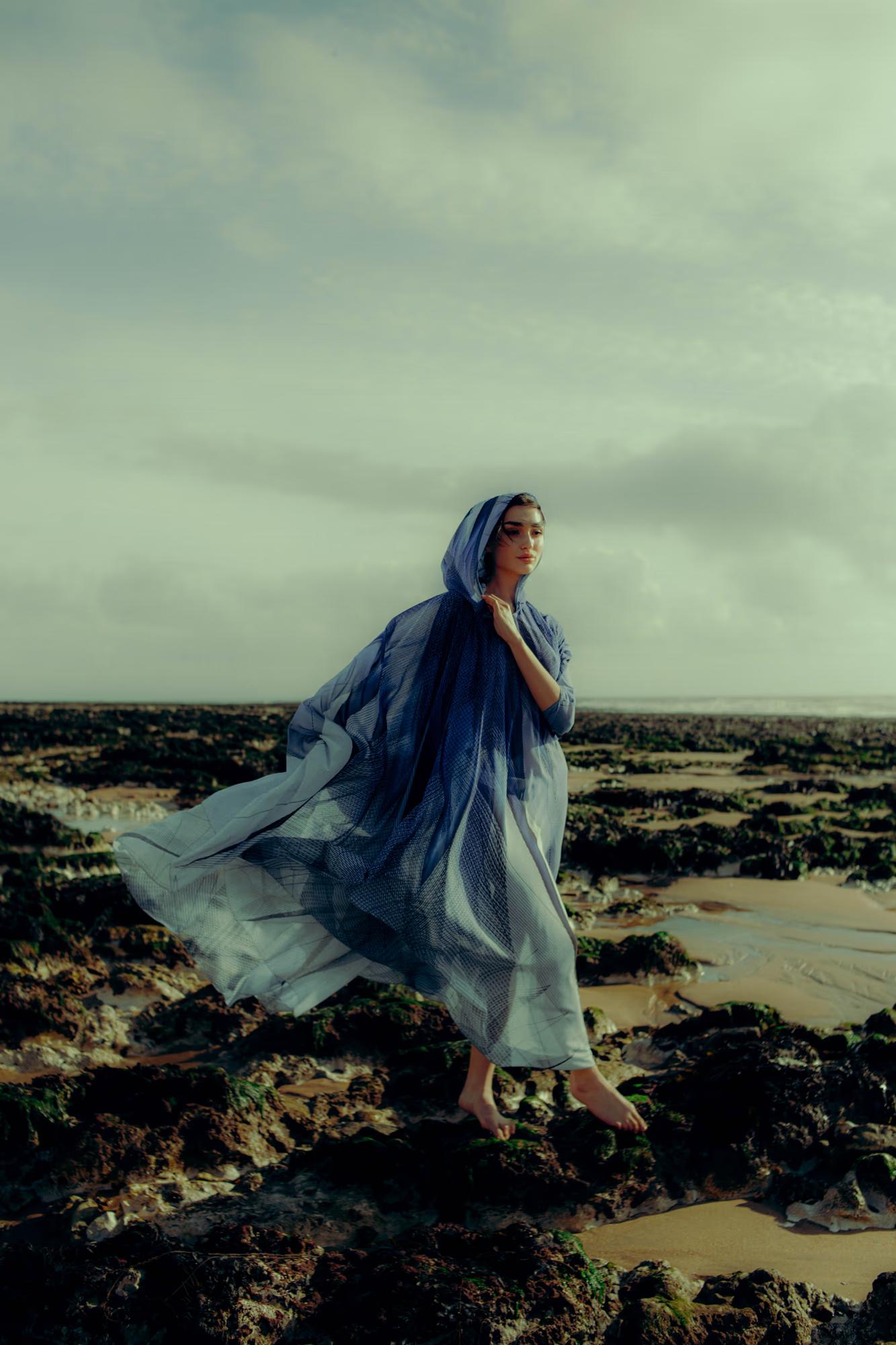 Full body fashion campaign shot of woman in blue designer cape walking along Eastbourne rocky coastline, dramatic cloudy sky, luxury editorial
