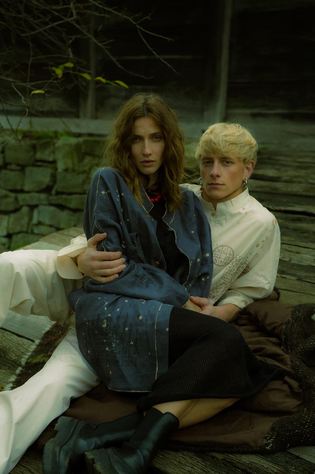 Cinematic fashion photography: models lounging in a field near authentic 19th-century rural architecture in Pirogovo museum.