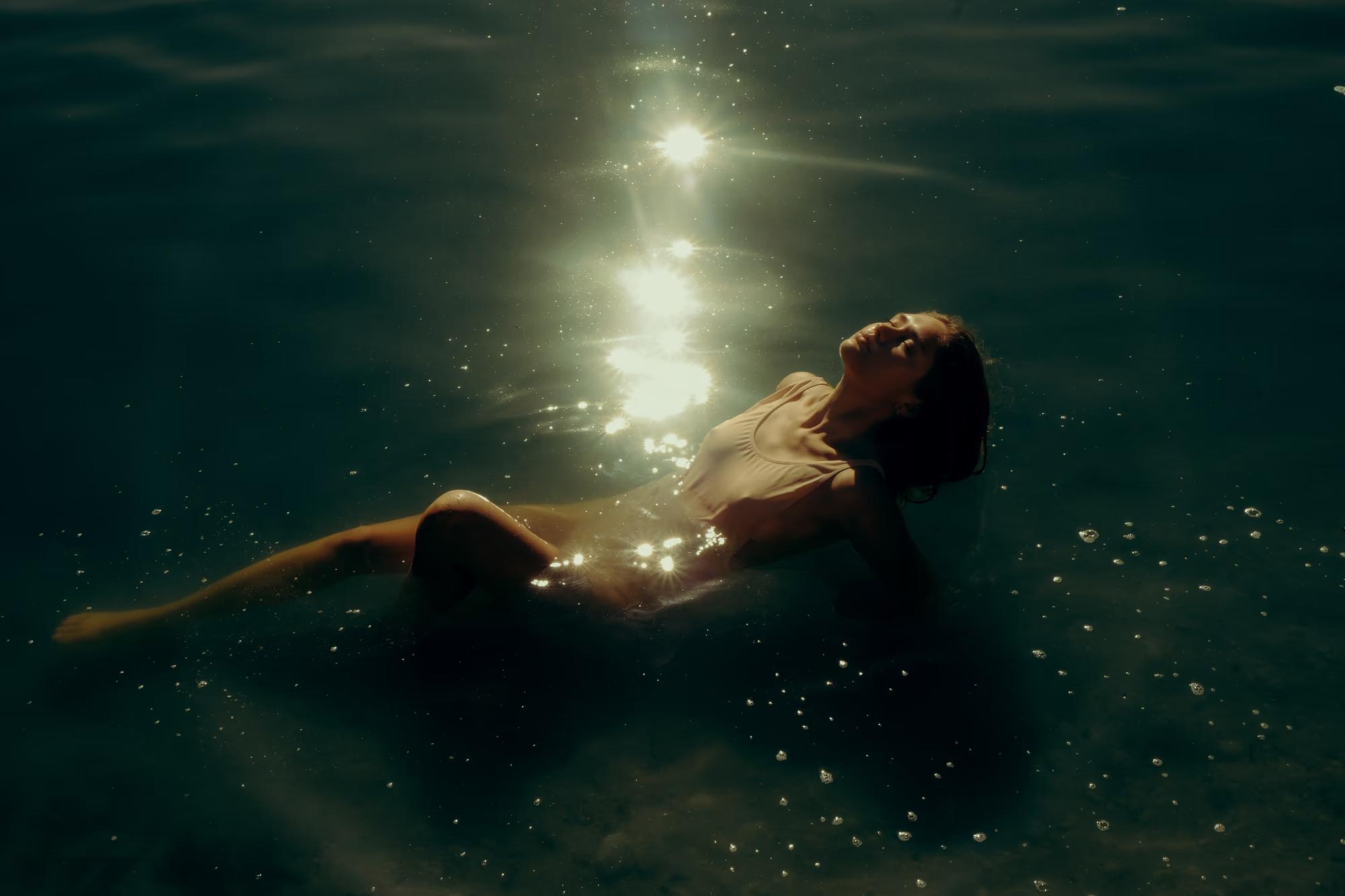 Underwater wide shot of woman floating near surface in dark lake with soft golden light reflection, ethereal cinematic fine art photography