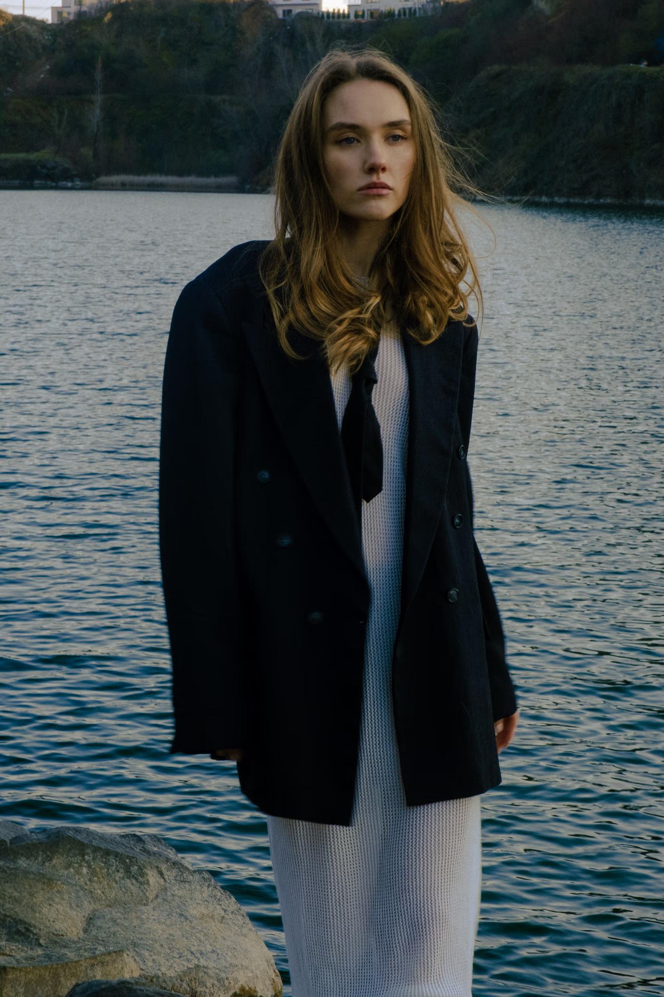 A fashion portrait showing a model with flowing hair wearing a dark blazer over a light slip dress, standing by the water's edge.