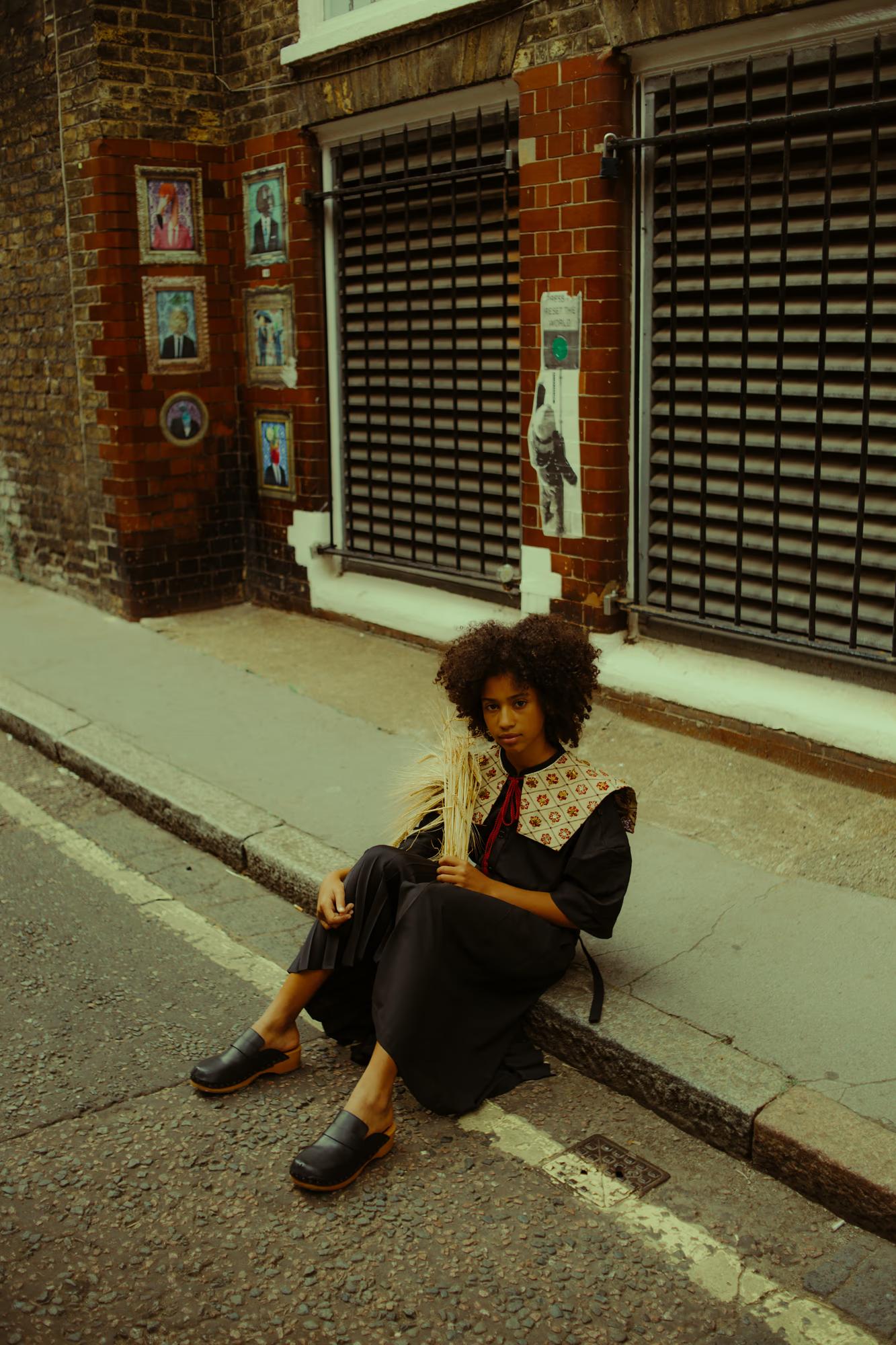 A model sits on a kerbstone in a London alley, wearing a black pleated skirt, an embroidered collar top, and chunky black wooden clogs. She holds a sheaf of dried wheat and looks directly at the camera. Behind her, a red brick wall is decorated with a cluster of framed surrealist paintings in gilded frames.