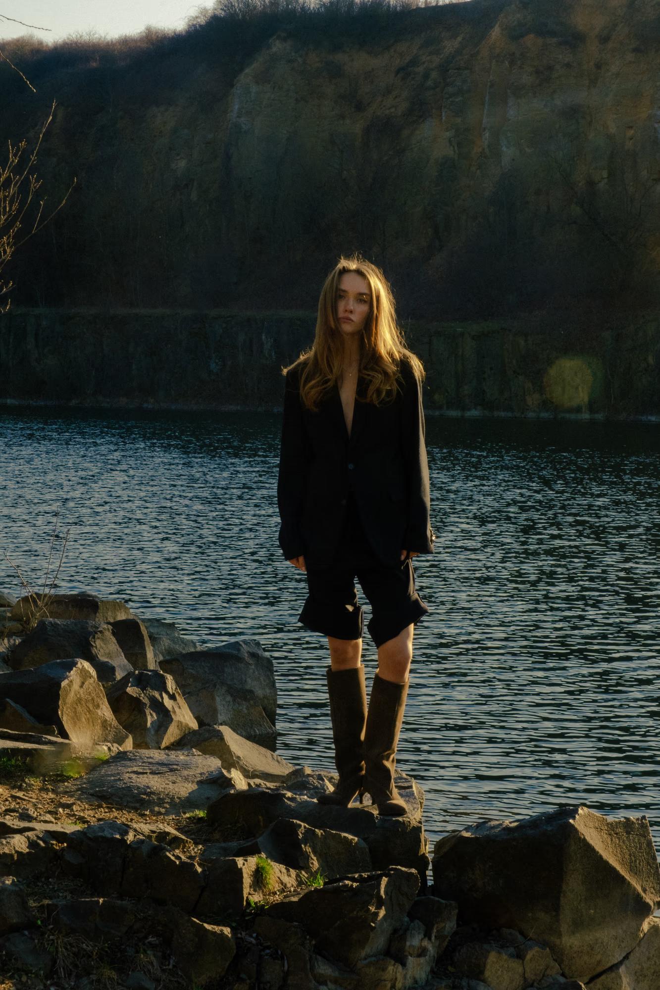 Wide-angle landscape shot: The model posing on the rocky shore of a large lake, showing the vast water and natural surroundings.