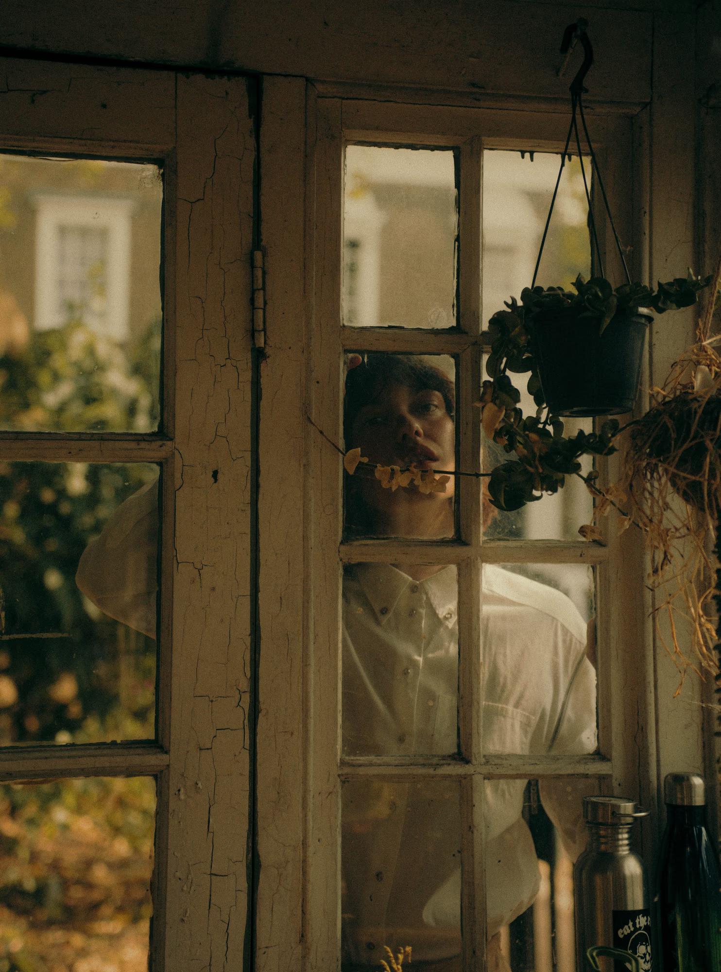 A model peers through the cracked, peeling panes of an old greenhouse window, face partially visible between the wooden mullions. She holds a small sprig of dried flowers to her lips and looks upward. A hanging pot plant and dried foliage are visible nearby. Warm amber light fills the frame.