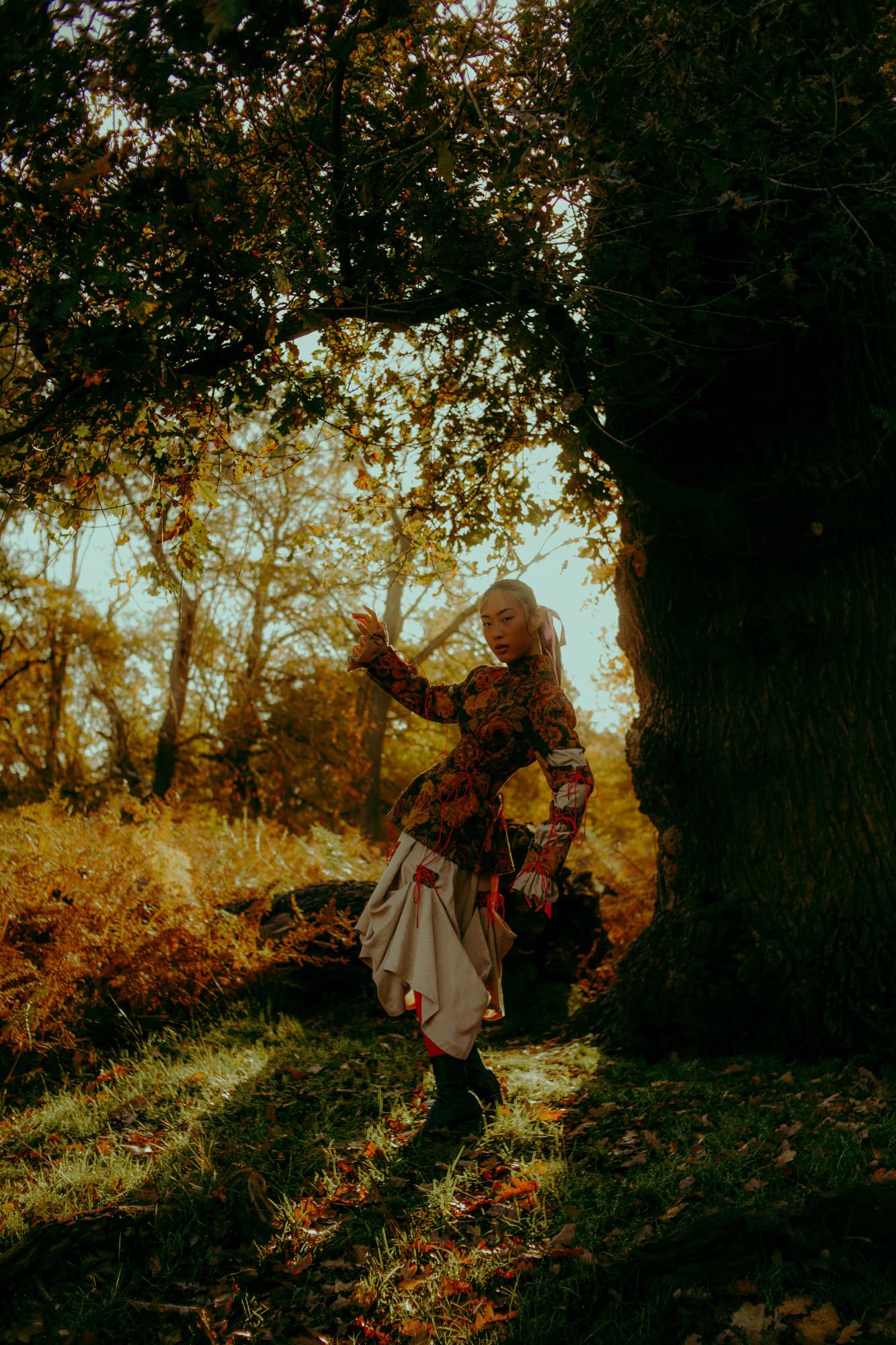 Medium fashion campaign shot of woman in designer floral outfit in Richmond Park London, soft golden hour light filtering through trees