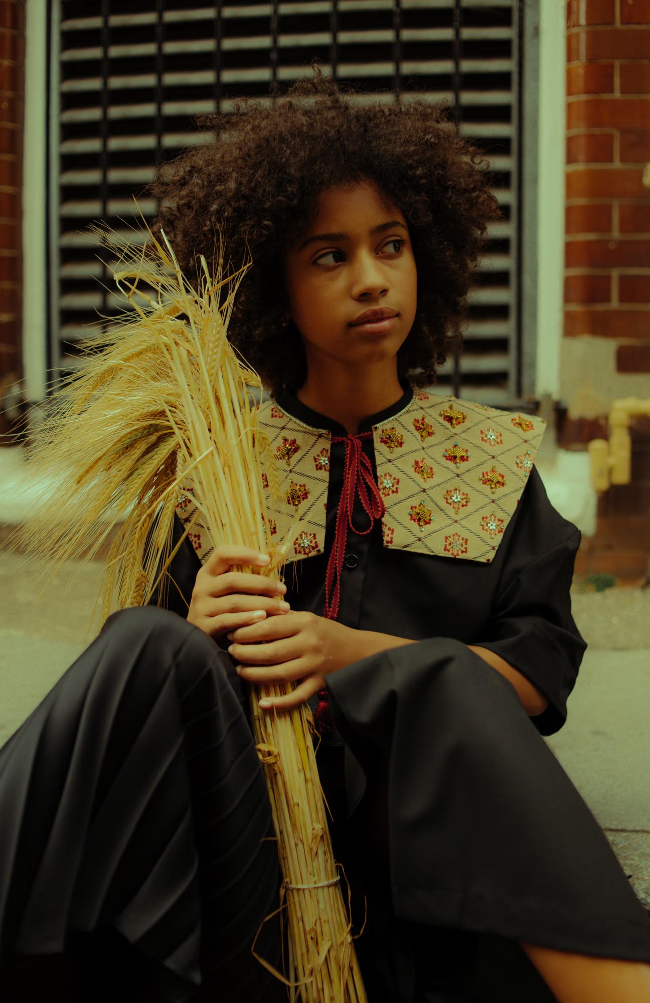Close-up portrait of a model seated on a pavement, holding a large bundle of dried wheat stalks. She wears a black coat with a wide embroidered diamond-patterned collar decorated with floral beaded motifs and tied with a red cord. Her natural curly hair frames her face as she gazes to the side.