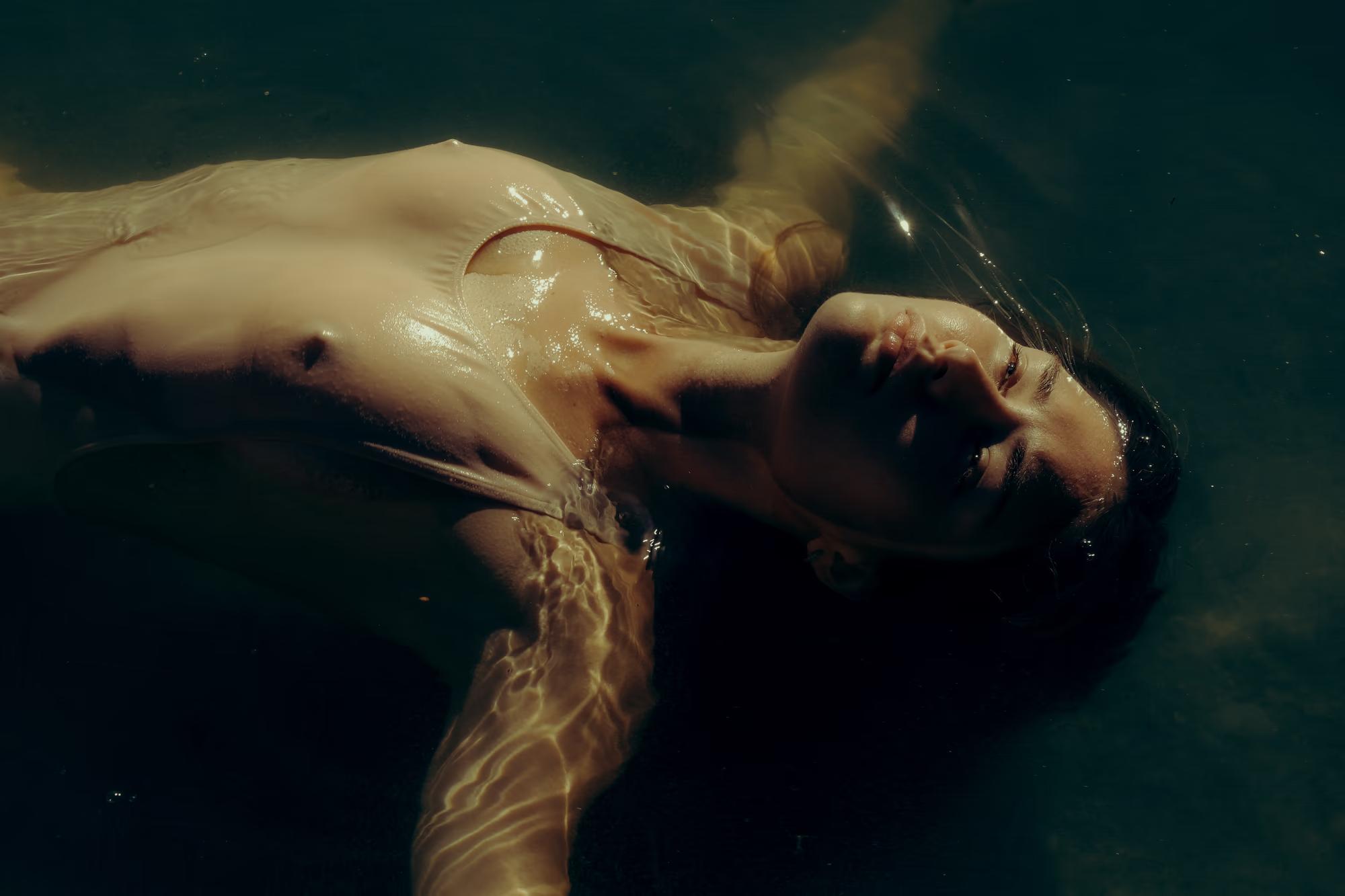 Underwater medium shot of woman floating face down in dark teal lake, long hair spread in water, cinematic underwater photography