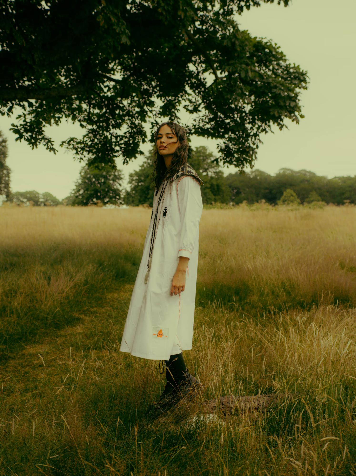 A model stands in profile on a fallen log in a sunlit meadow, wearing a long white dress with folk embroidery along the yoke and cuffs, black figurine motifs on the fabric, and a long braided cord necklace with a pendant. She wears tall black boots and looks to the side. A large tree and golden grass fill the background.