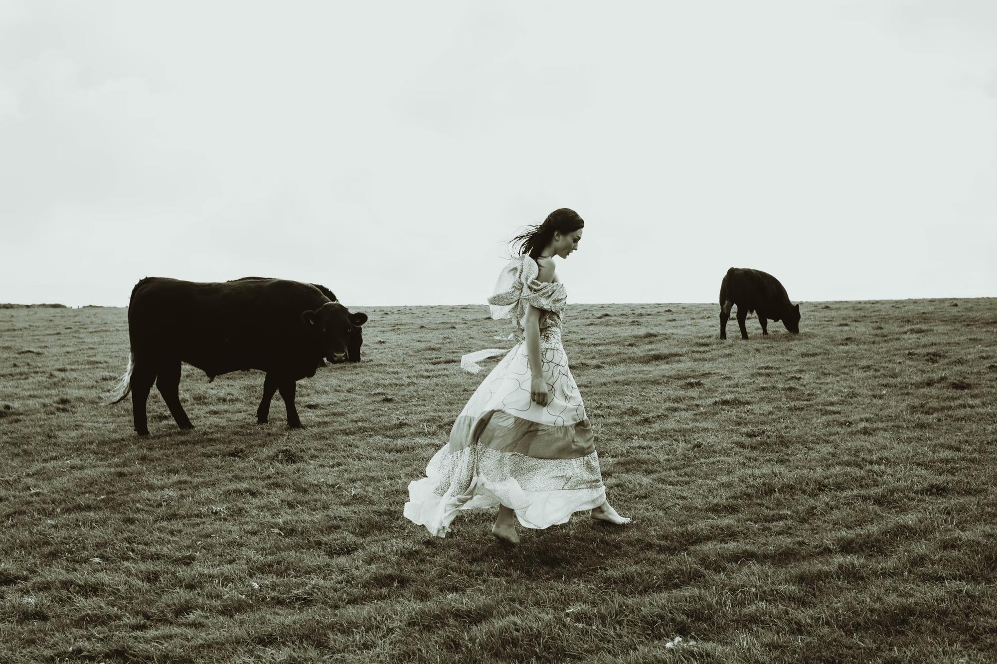 Full body fashion campaign shot of woman in white flowing designer dress on Eastbourne beach with black cow in background, editorial luxury fashion