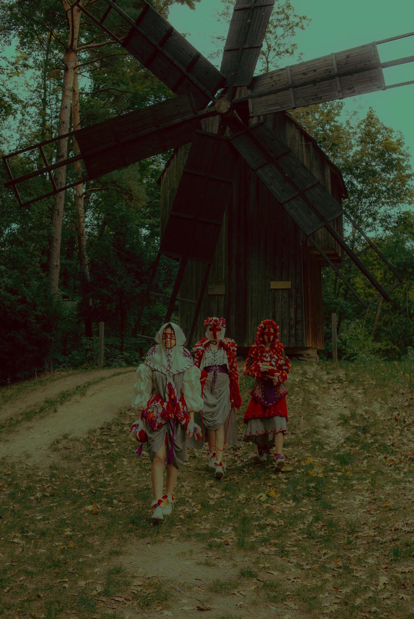 A wide-angle artistic shot of models in ethnic clothing walking towards an old wooden windmill, blending fashion with Ukrainian rural heritage.