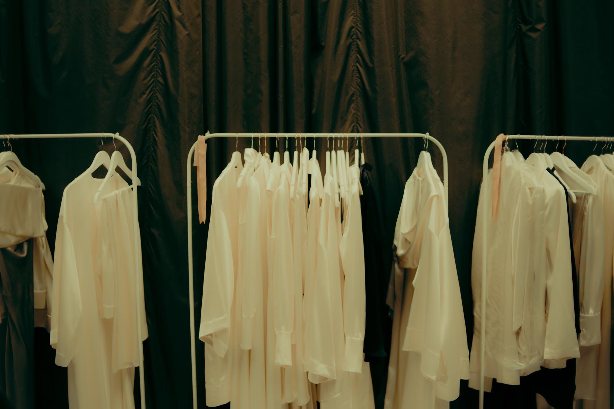 A rack of elegant white designer garments on hangers, ready for the models at a London Fashion Week show.