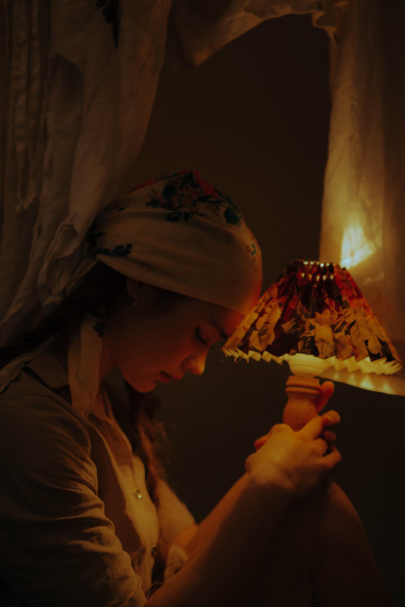 Medium fashion art shot of woman in traditional white outfit leaning over candles on floor, warm golden candlelight, dark atmospheric interior, ethno art