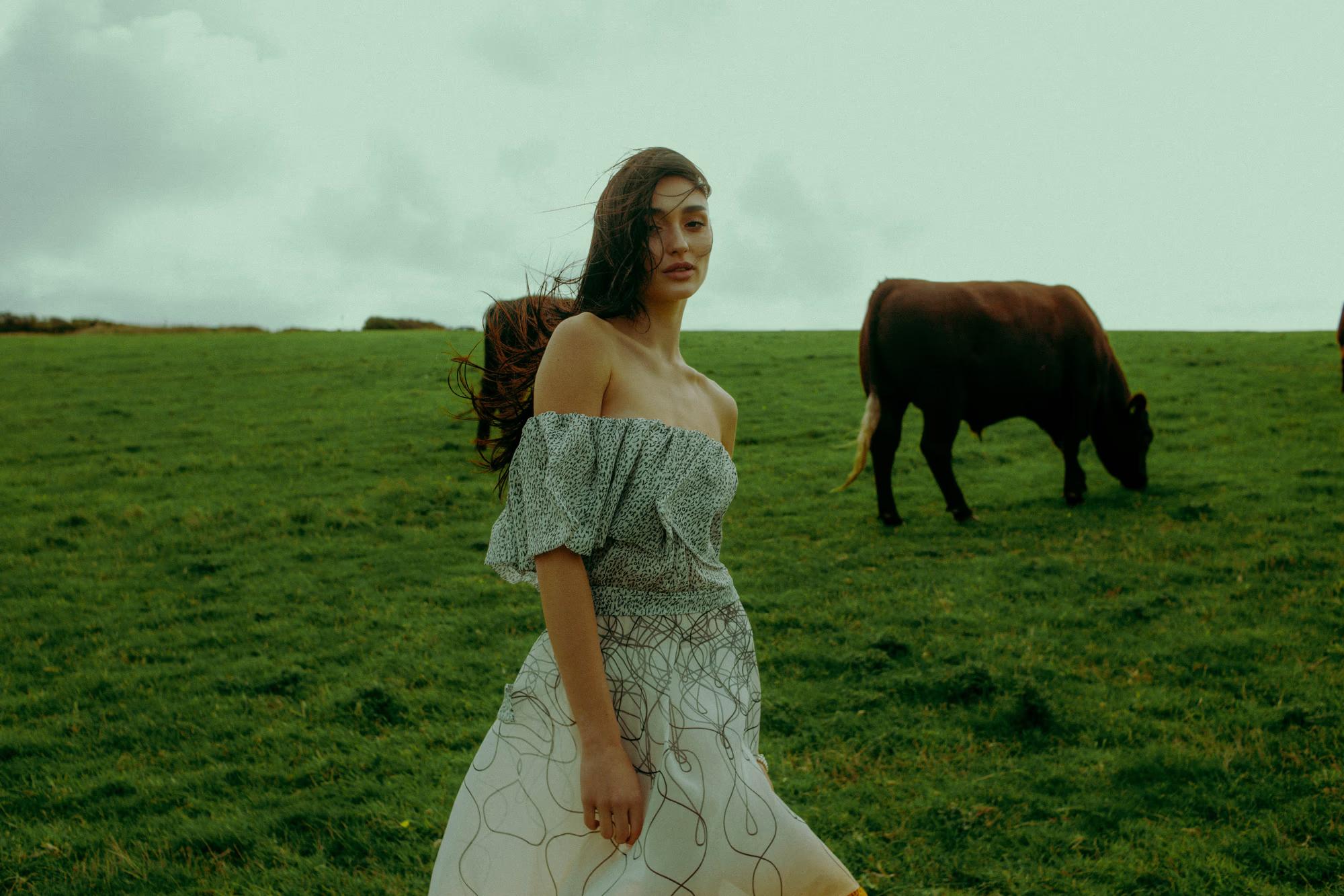 Full body fashion campaign shot of woman in white flowing designer dress standing in green coastal field with black cow grazing nearby, Eastbourne luxury editorial