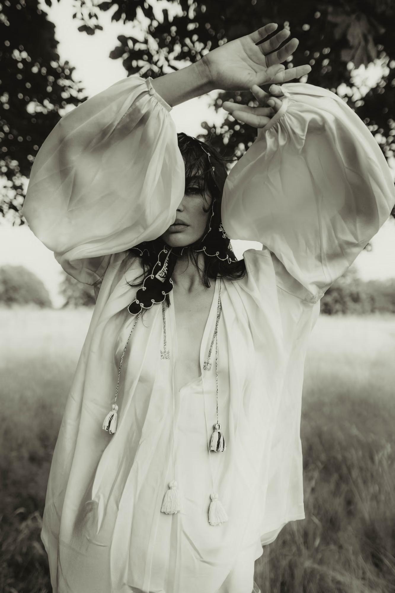 Black-and-white photograph. A model raises both arms overhead with hands crossed, wearing a voluminous white dress with dramatically wide puff sleeves. Around her neck sits a decorative dark collar with embroidery and dangling tassel ties. Her face is turned to the side with eyes closed. Blurred meadow and trees fill the background.