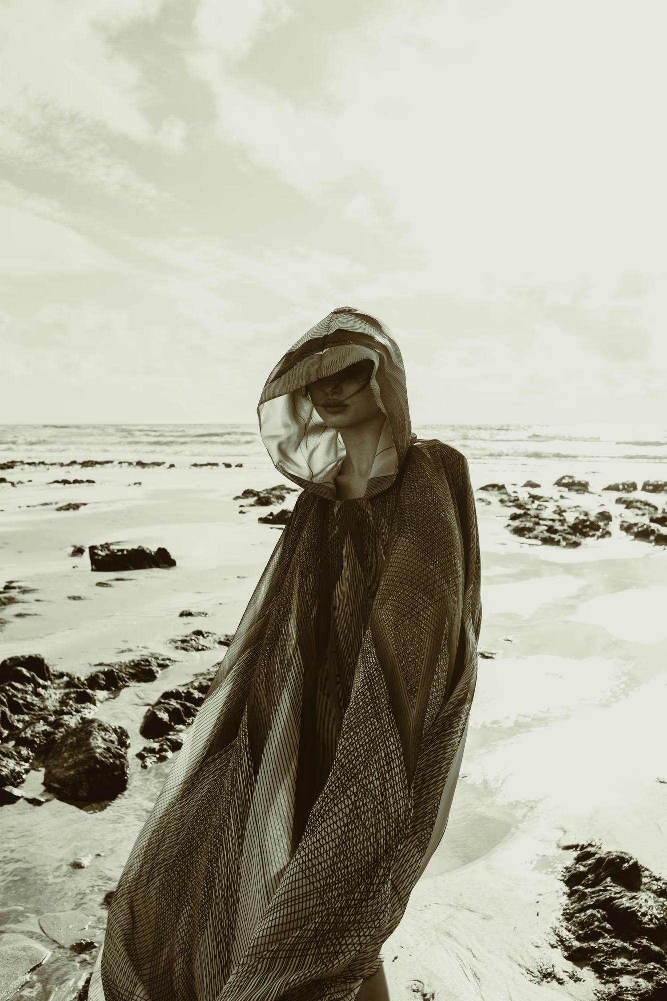 Full body fashion campaign shot of woman in oversized beige designer coat on rocky Eastbourne beach, windswept coastal editorial, luxury fashion