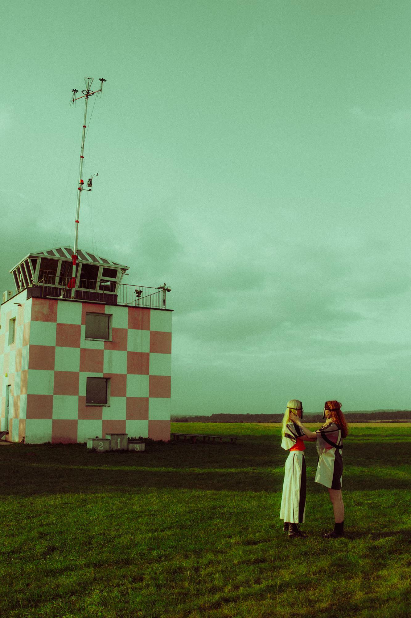 Wide shot of two masked models standing close together on a large grassy airfield, facing each other. Behind them rises a vintage red-and-white checkered airfield control tower fitted with a wind vane mast. The landscape is flat and open under a moody teal sky.