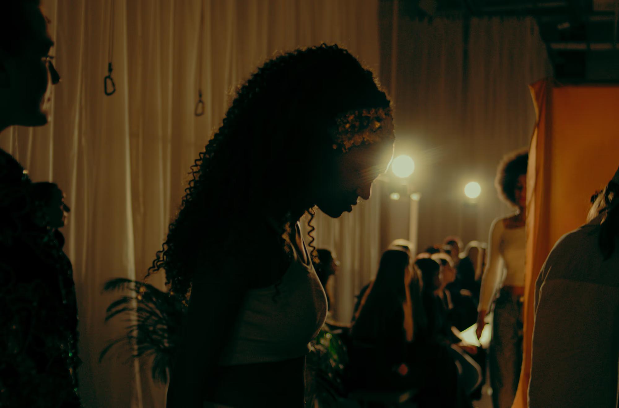 Silhouette of a model waiting in the wings of the fashion show, dramatic warm lighting highlighting the backstage atmosphere.