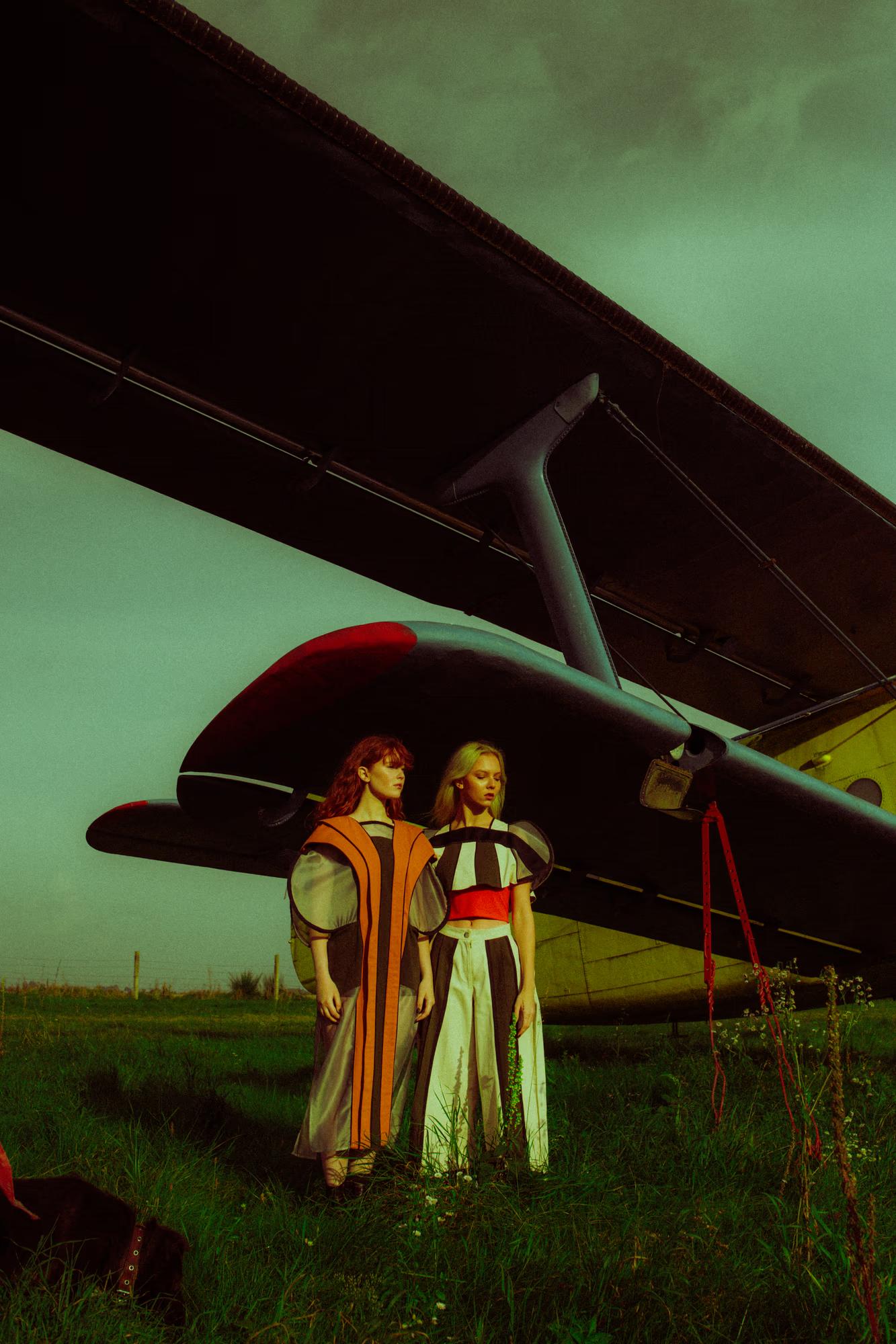  Two models stand together beneath the low-swept wing of a dark vintage aircraft on a grass field at dusk. The redheaded model wears a sheer amber-panelled cape; the blonde model wears a bold red, black, and white structured top with wide-leg striped trousers. A stormy teal sky looms overhead.