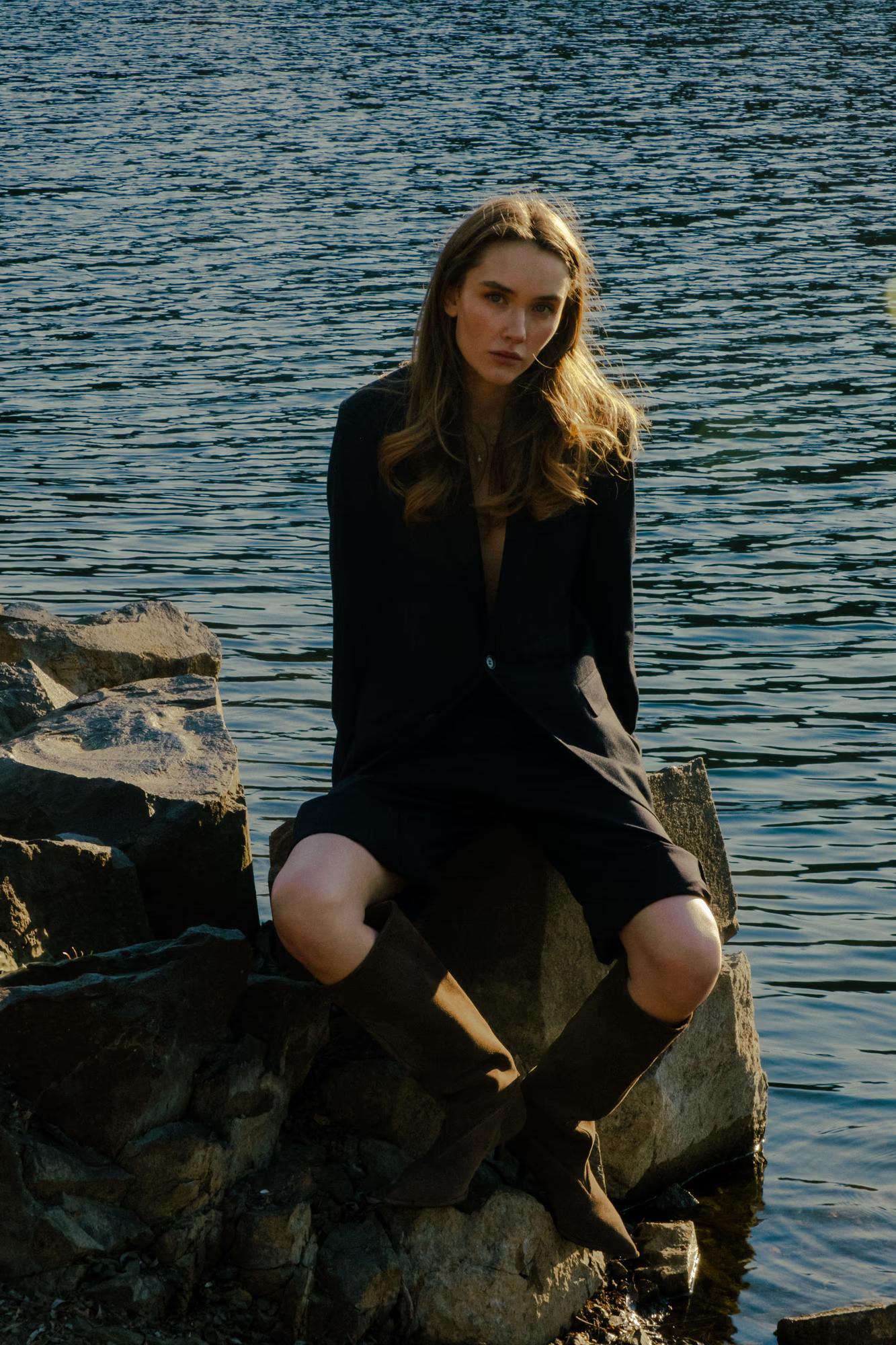 Distant shot: The model sits high on a rock formation above the lake, framed by the landscape at twilight.
